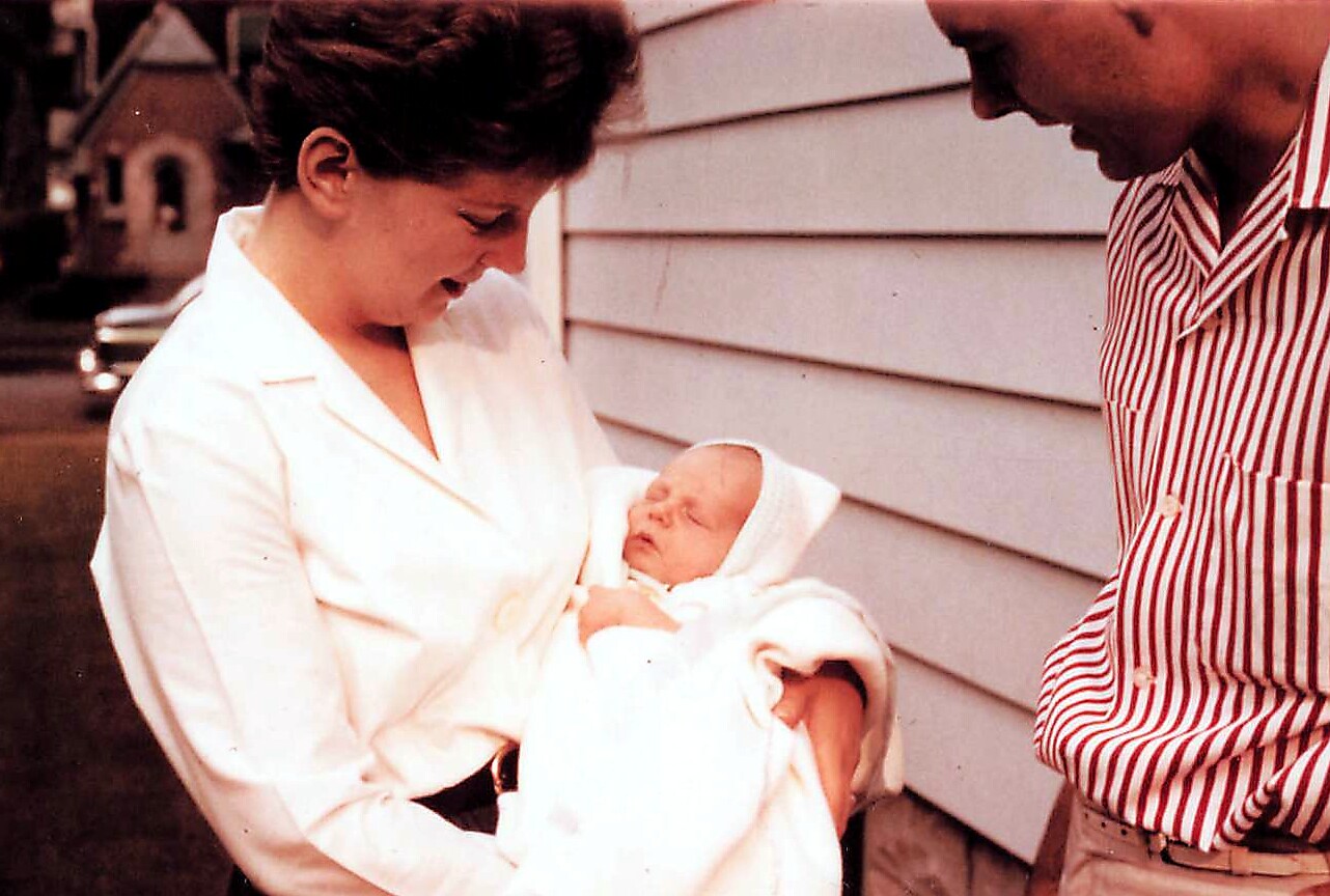 Woman in a white blouse cradles a sleeping newborn wrapped in a blanket, outdoors beside a house as a man in a striped shirt looks on.