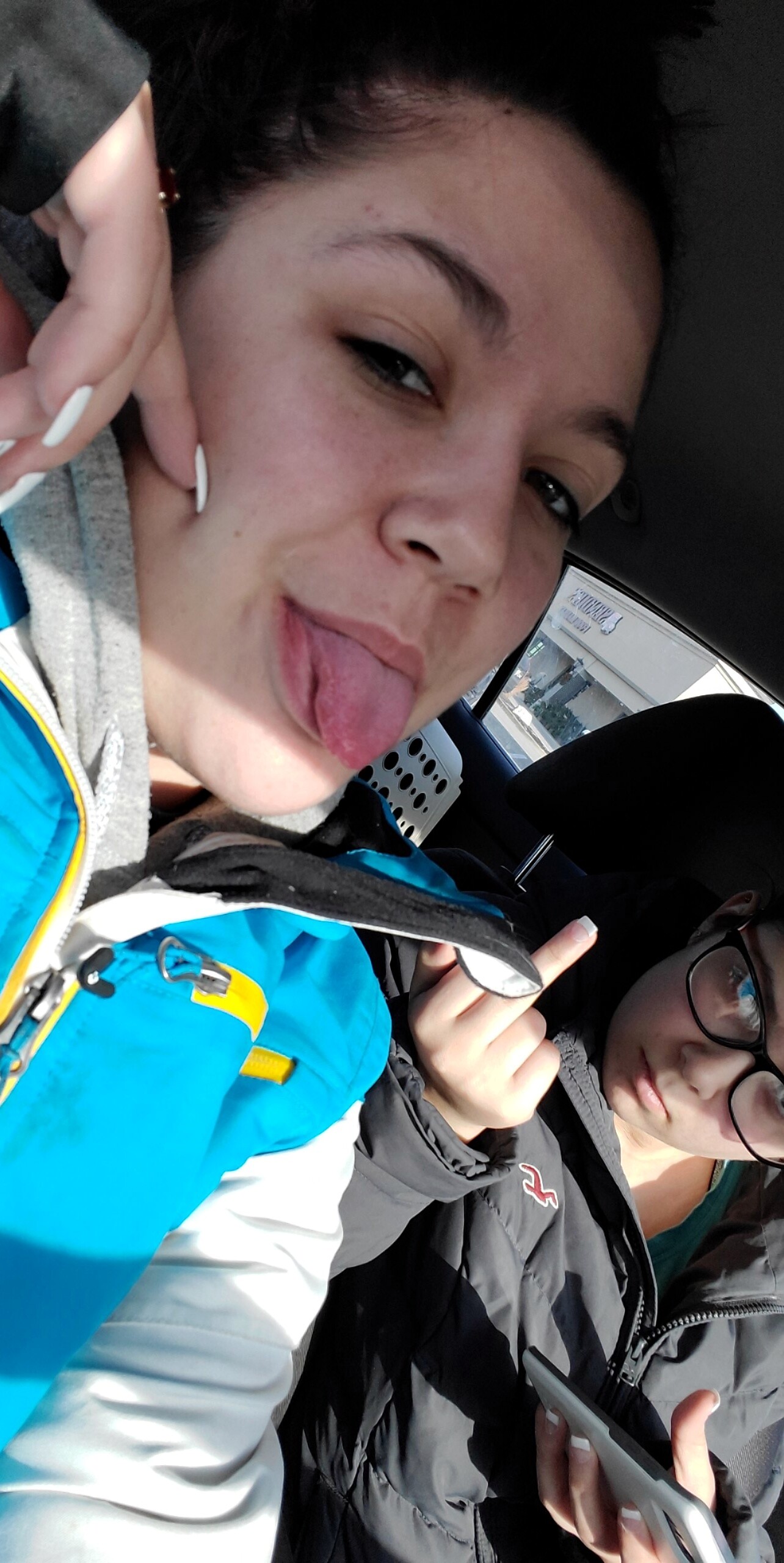 Two people in a car, the person in front sticking out their tongue playfully at the camera while the passenger wears glasses and looks sideways.