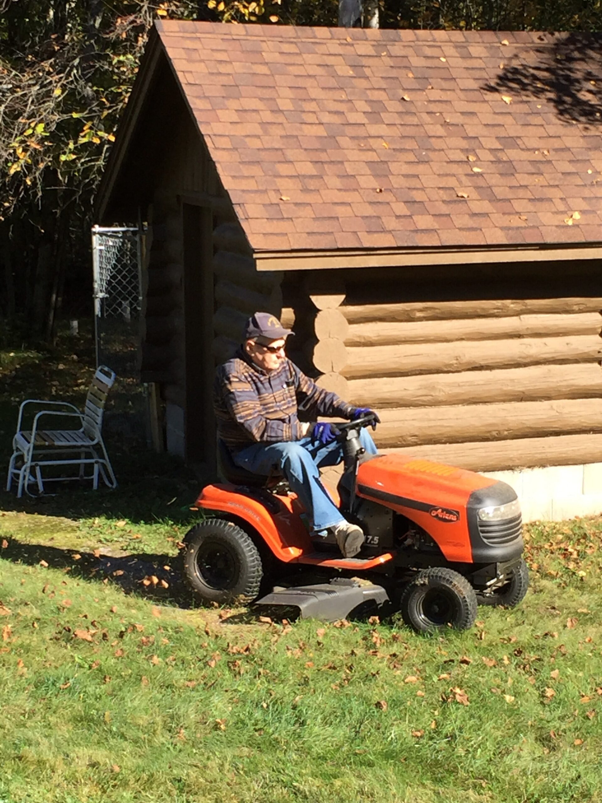 Older man wearing a cap rides an orange riding lawn mower in a grassy yard in front of a log cabin under autumn light.