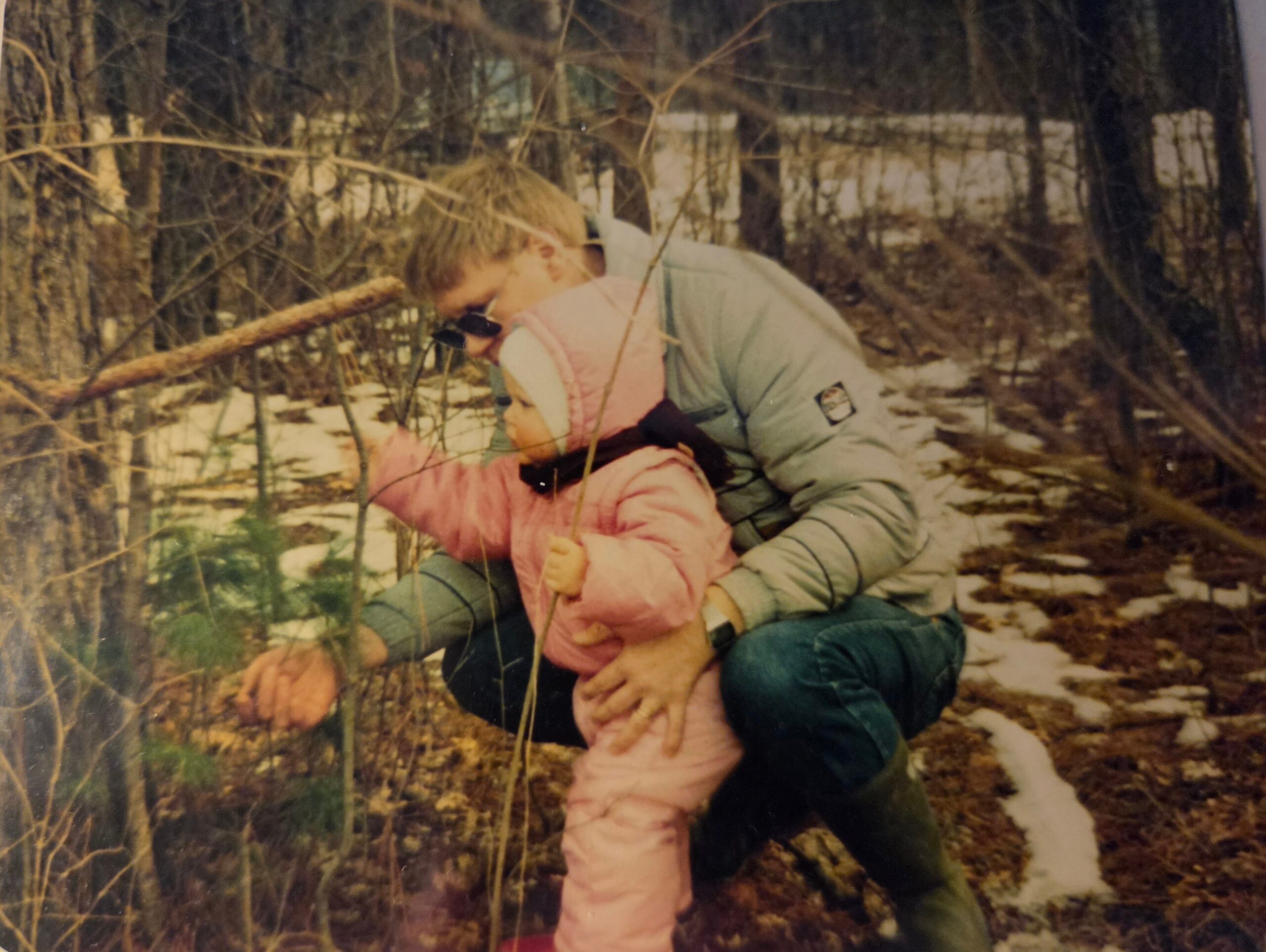 Man in a light jacket supports a child in a pink snowsuit outdoors in a snowy, wooded area.