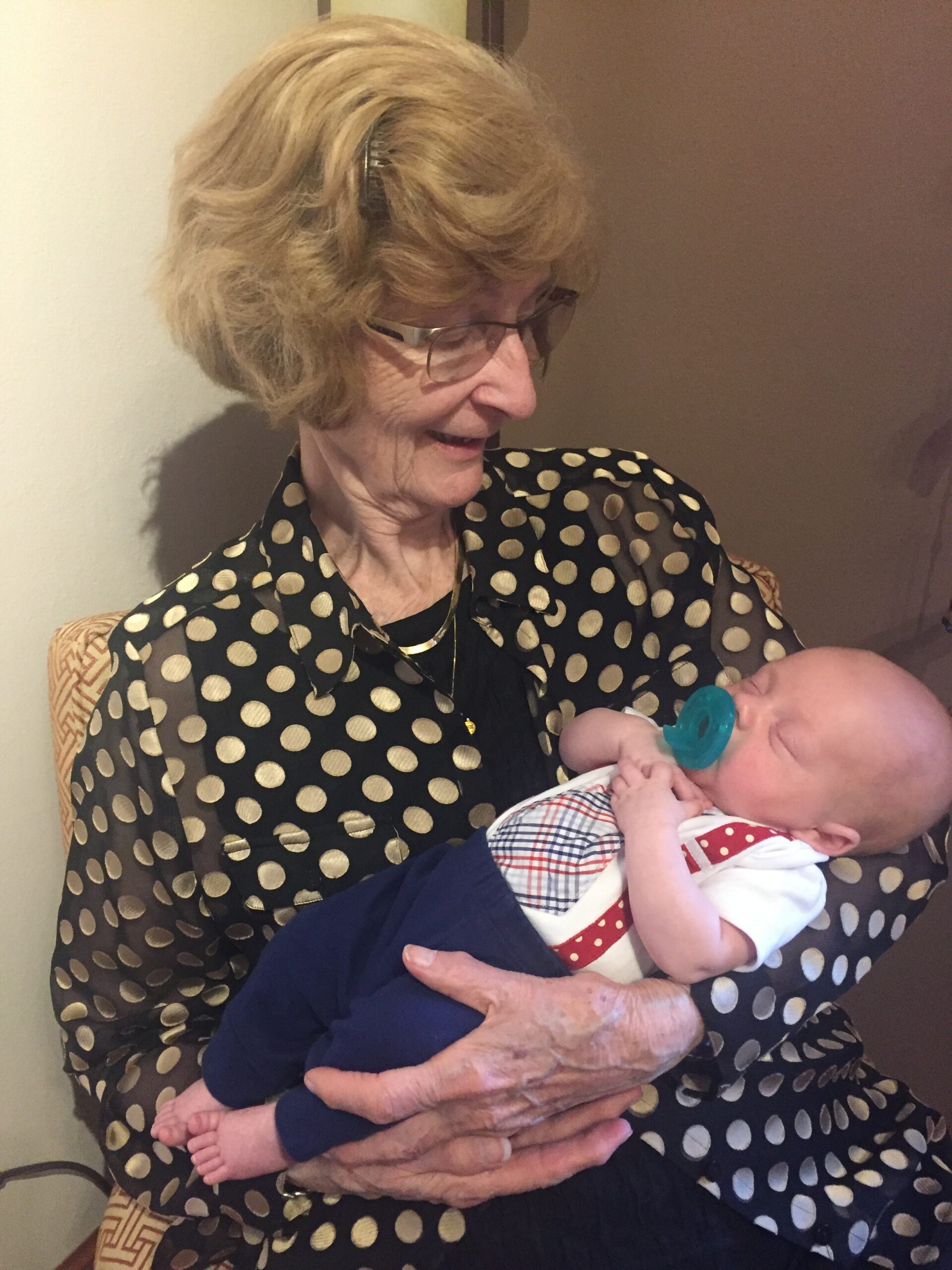 Older woman with short blond hair and glasses cradling a newborn baby wrapped in a blue blanket and a teal pacifier in the baby's mouth.