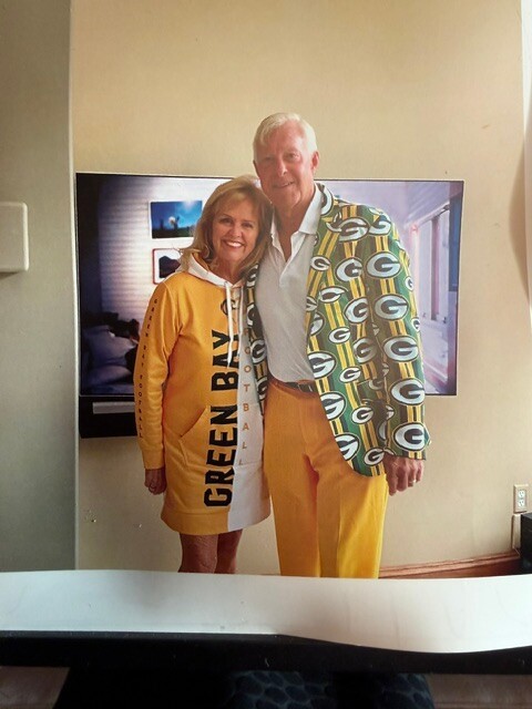 Smiling older couple posing indoors in bright yellow outfits: woman wearing a yellow hoodie-style dress with 'GREEN BAY' text along the side, man in a yellow suit with a bold green-and-white patterned jacket.