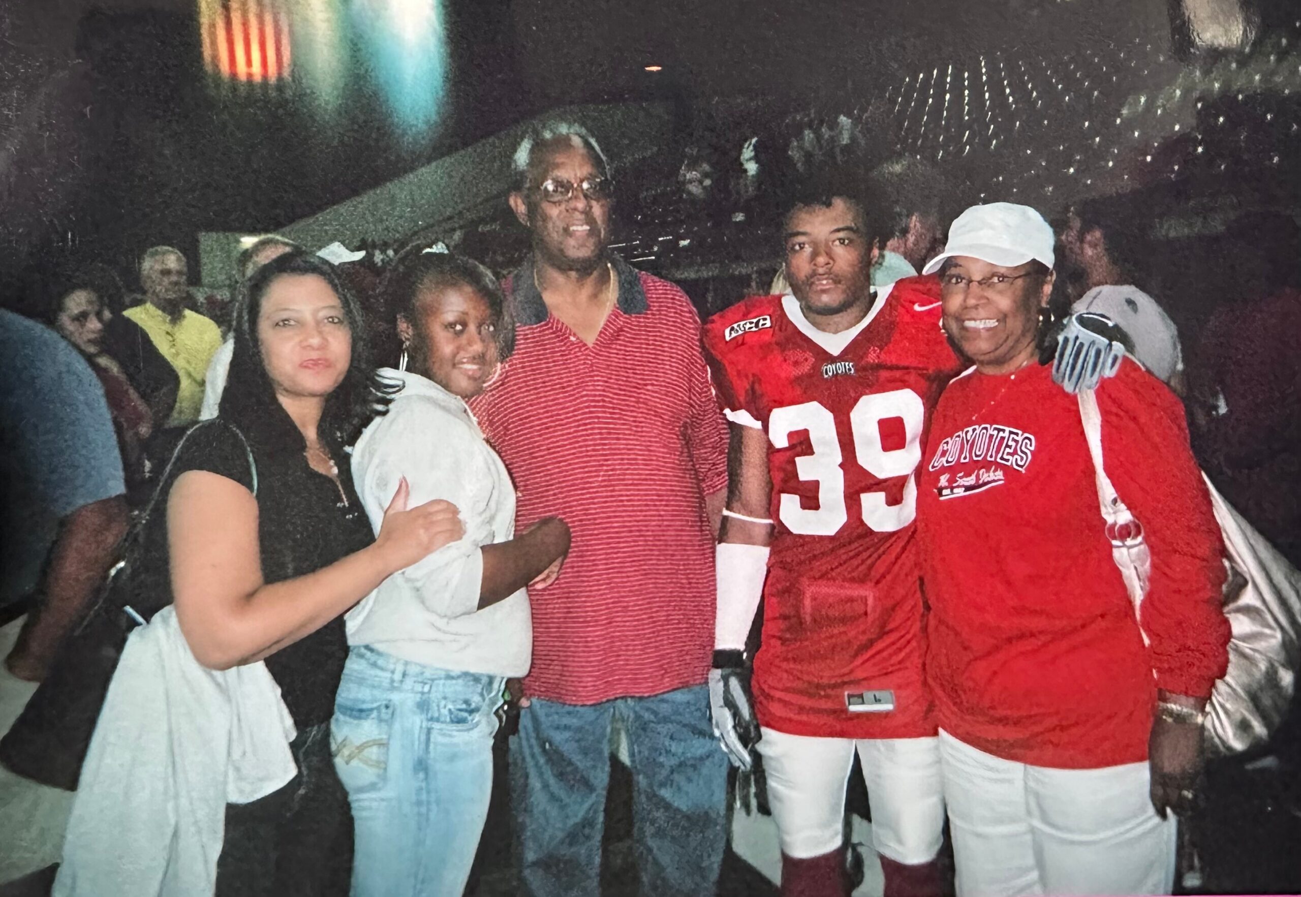 Group of five people posing together at night, two women in front left, two men in the middle, and a football player in a red Coyotes jersey on the right.