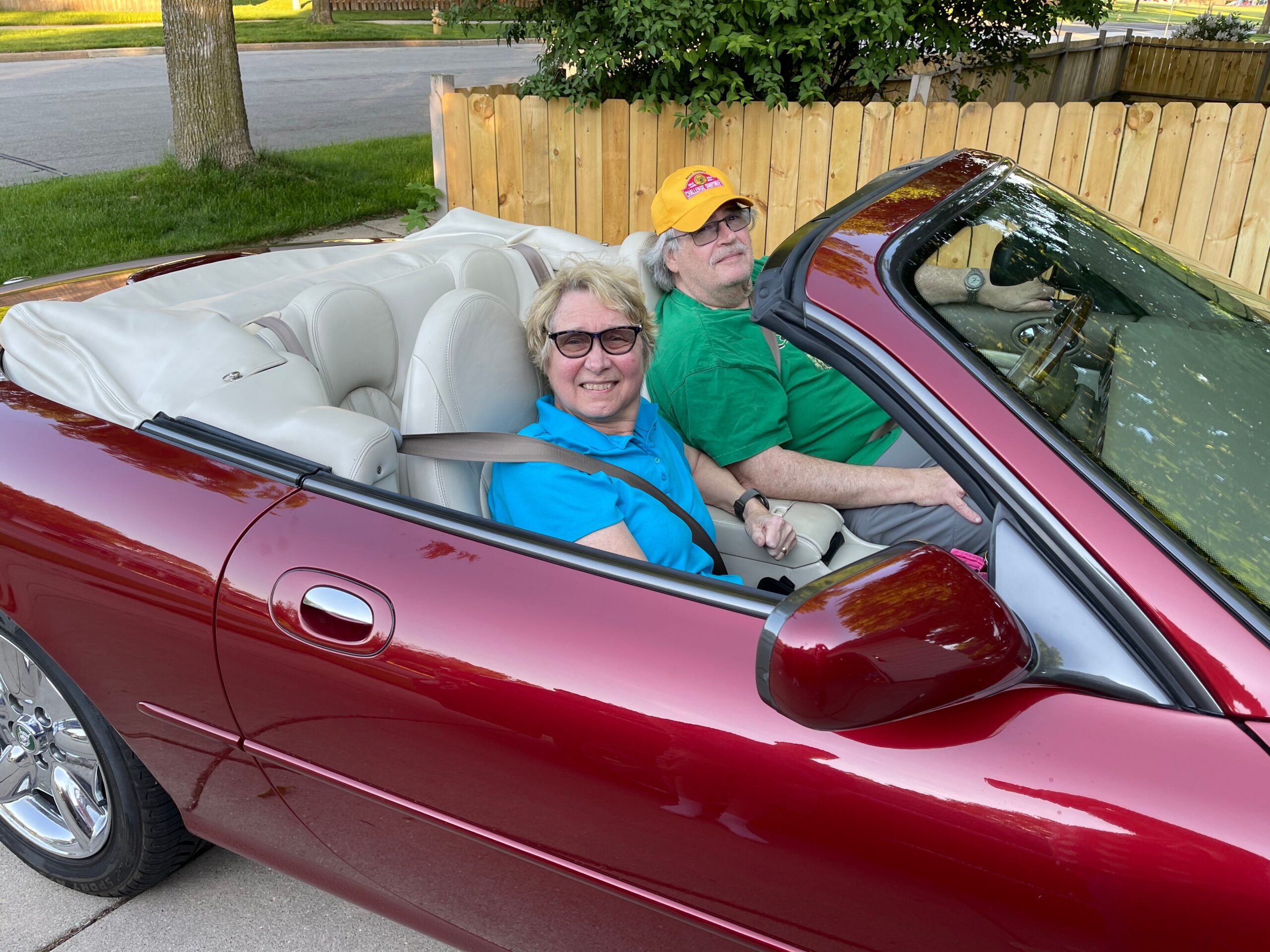 Smiling couple in a red convertible with white interior parked on a suburban street; woman in blue in the passenger seat, man driving in green shirt and sunglasses.