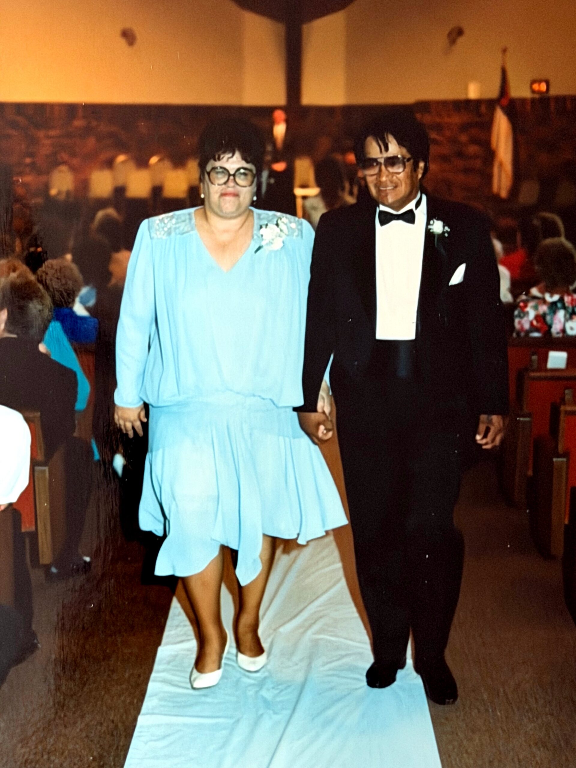 Couple walking hand-in-hand down an aisle at a formal event; woman in a light blue dress with pearl accessories, man in a black tuxedo with bow tie.