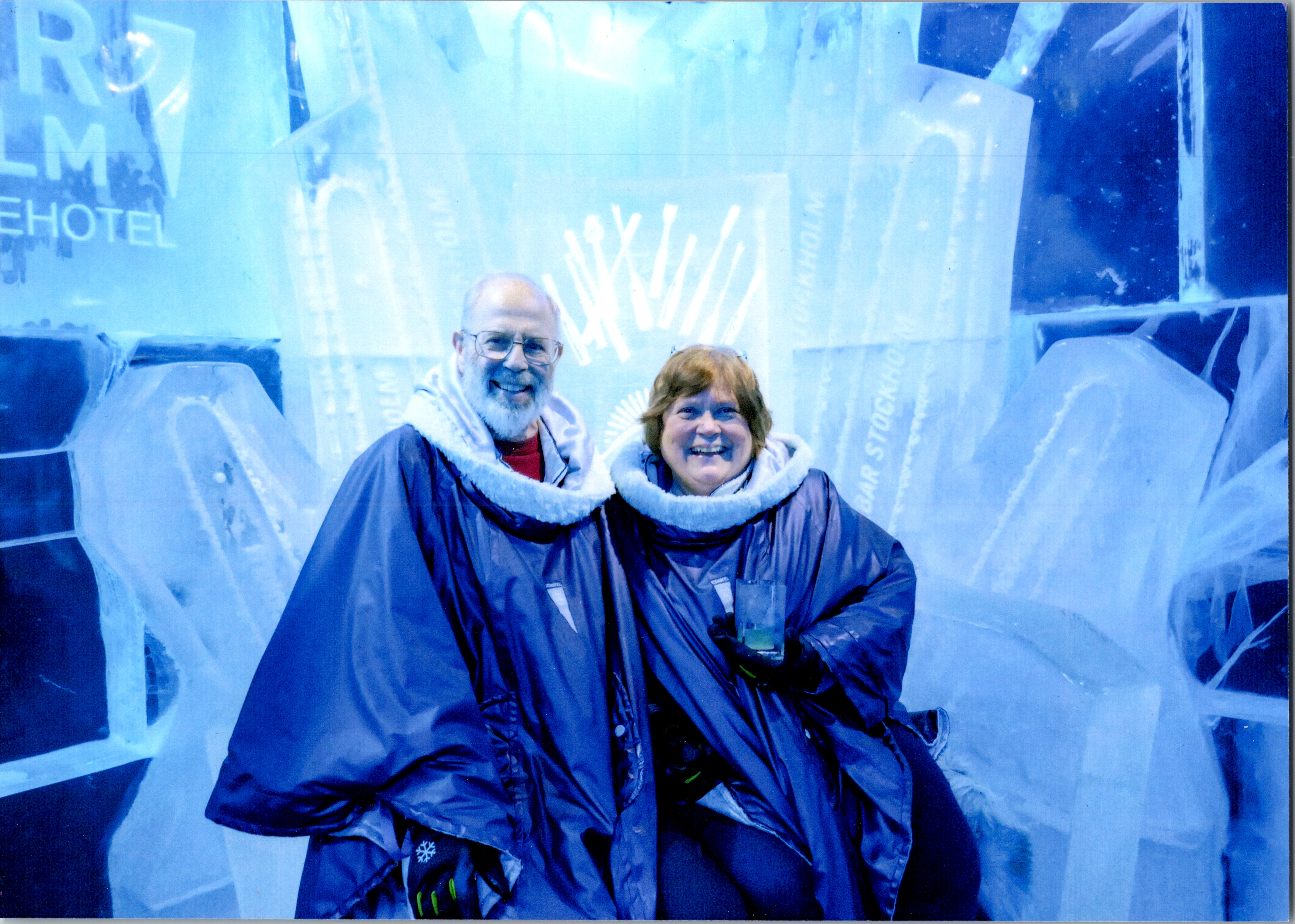 Two smiling adults in matching blue winter parkas posing in front of an ice sculpture backdrop.