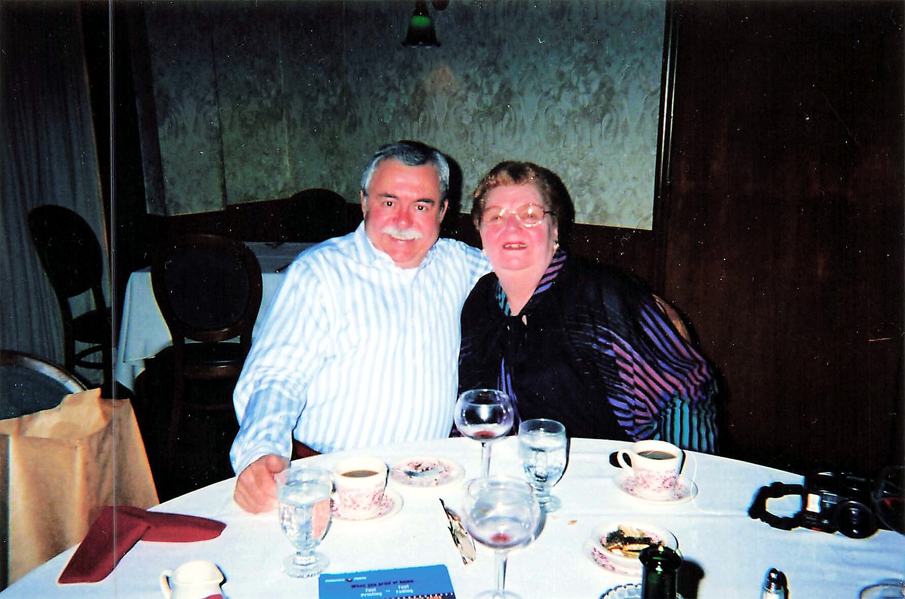 Older couple smiling at a restaurant table with coffee cups, wine glasses, and desserts in front of them.
