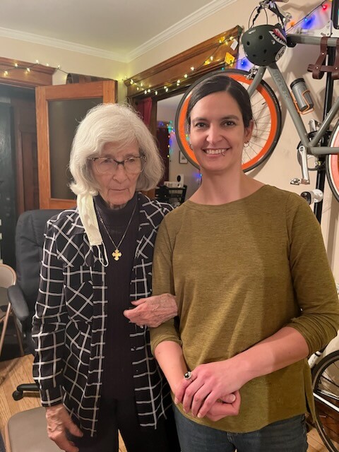 Two women posing indoors: an elderly woman with white hair and glasses in a checkered jacket, and a smiling younger woman in a mustard top.