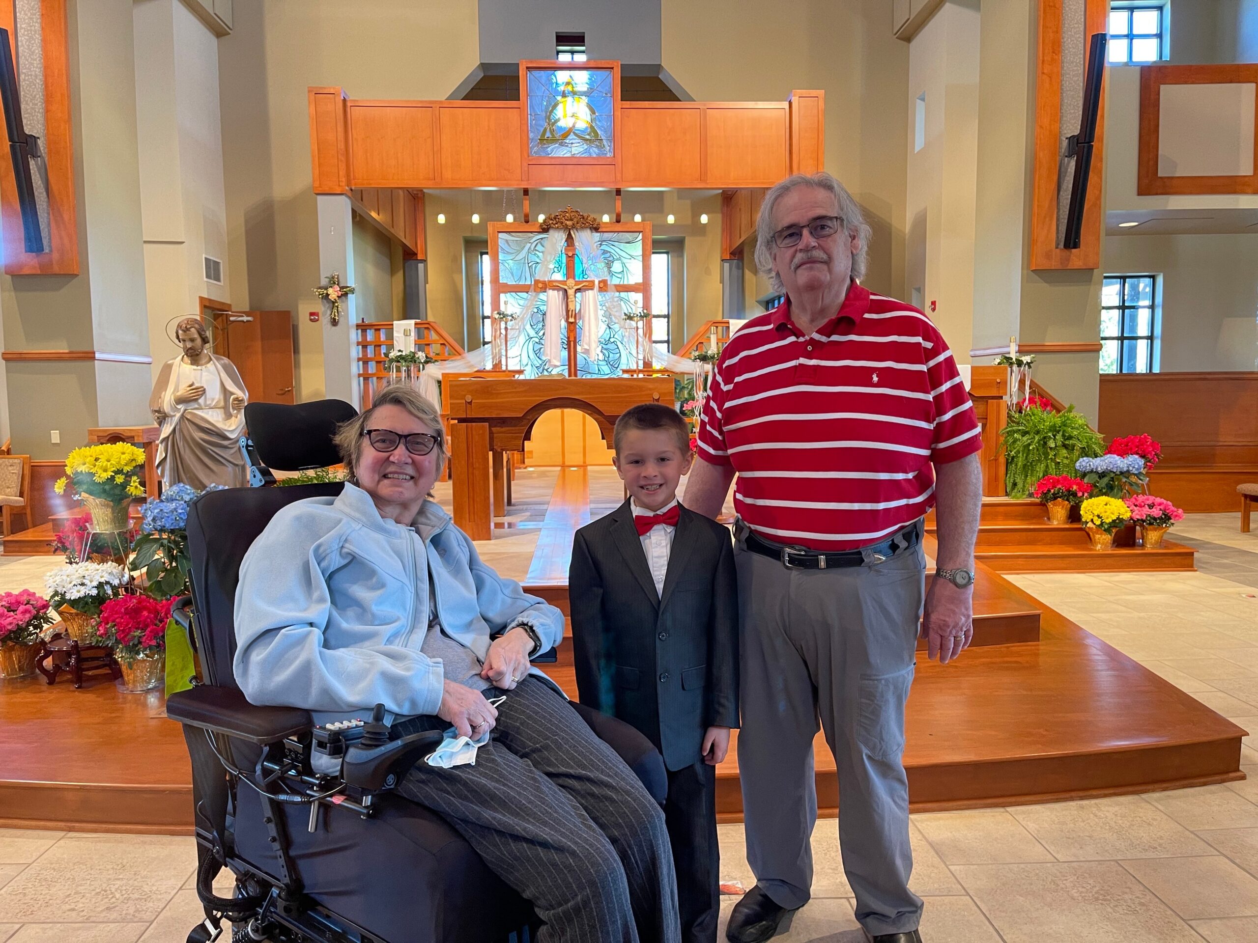 Three people pose in a church sanctuary: an elderly woman in a wheelchair, a boy in a suit with a red bow tie, and a man in a red striped shirt standing near the altar with flowers and stained-glass windows behind them.