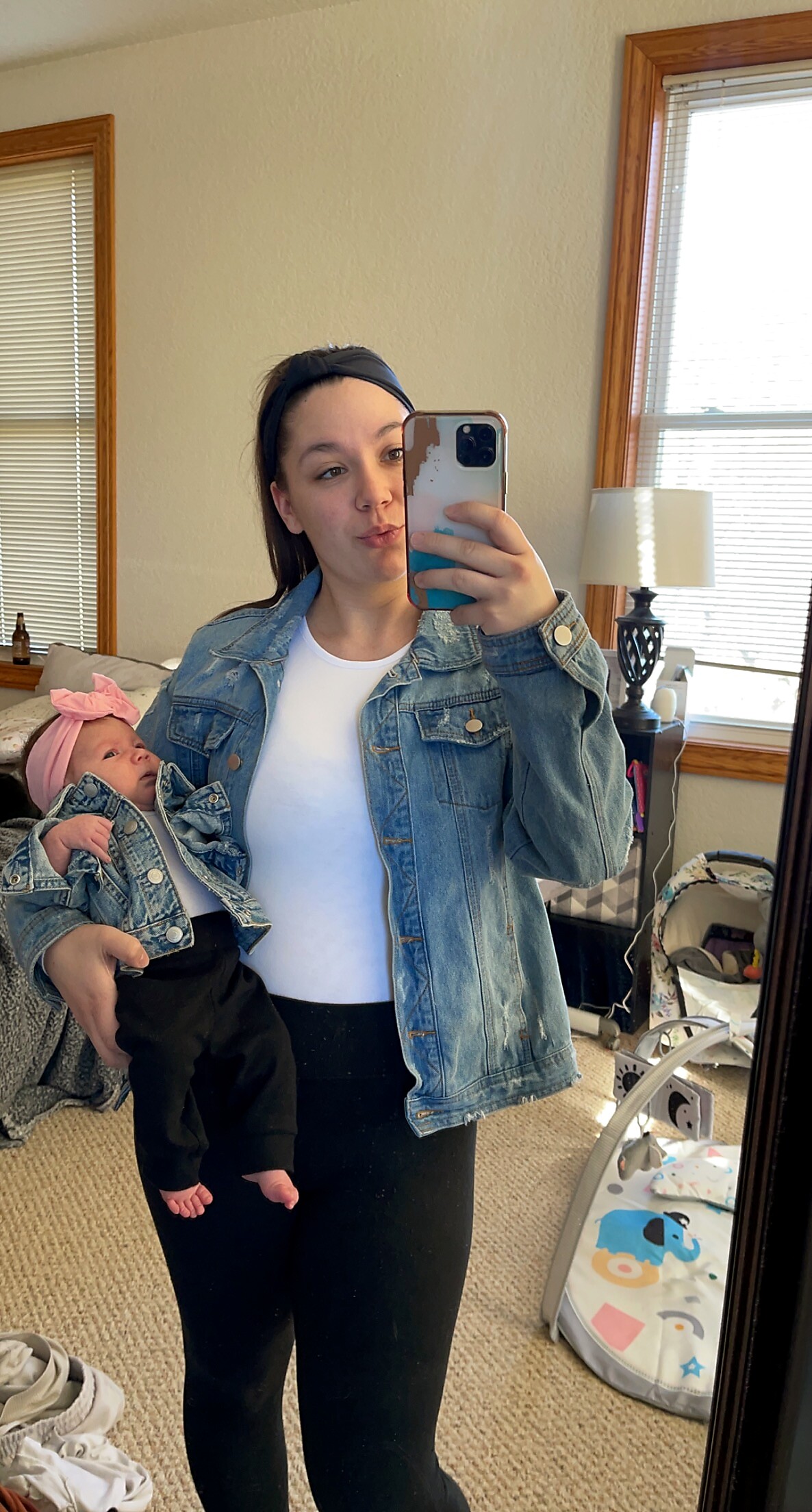 Young woman in a denim jacket taking a mirror selfie while holding a baby girl in a denim jacket and pink headband.