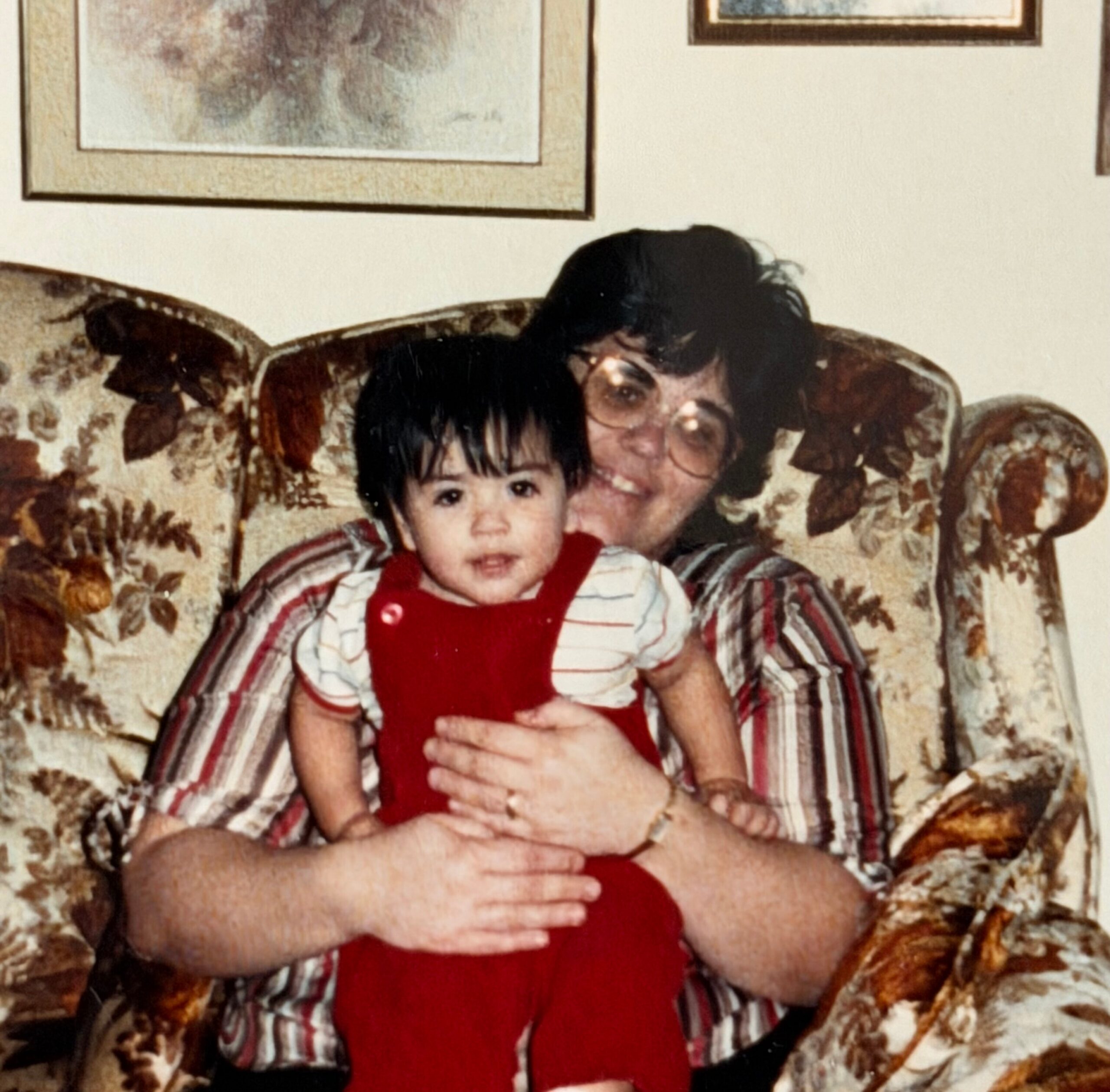 A young child in red overalls sits on a floral sofa, with an older child and adult nearby on the couch behind them.