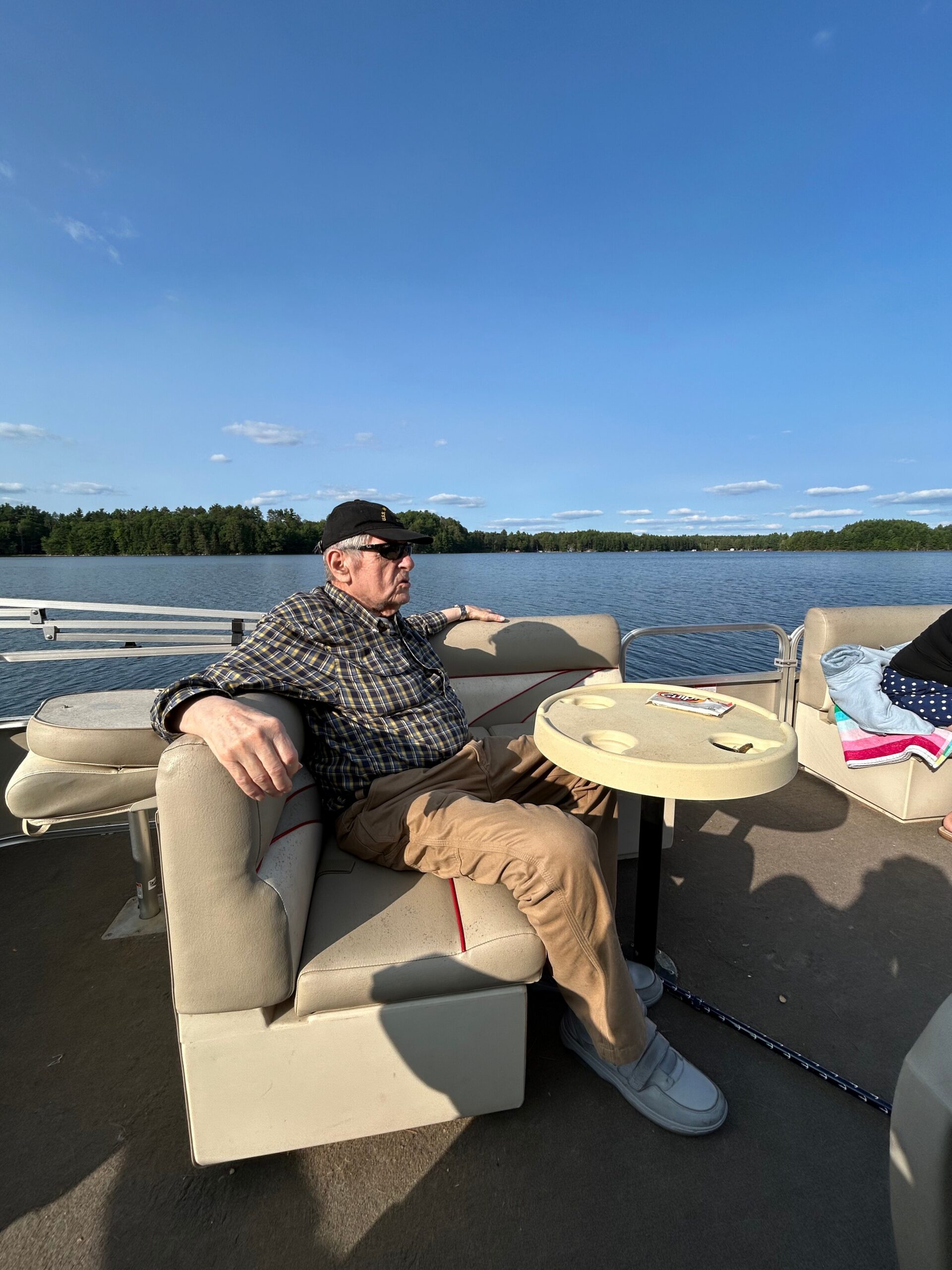 Older man wearing a black cap, sunglasses, and plaid shirt relaxing on a pontoon boat by a calm lake with trees in the distance.