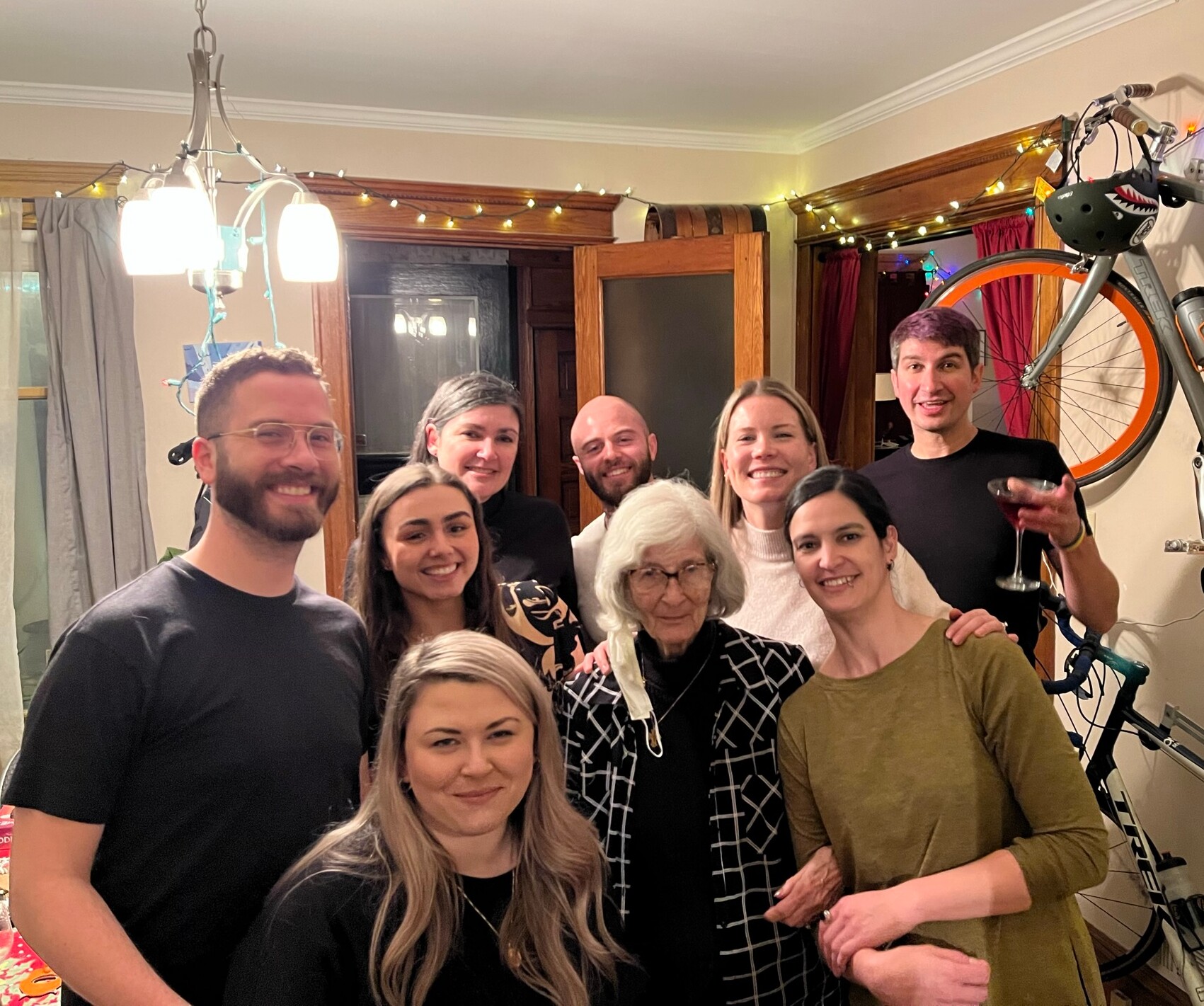 Group of smiling adults posing together in a cozy living room with string lights and a wall-mounted bike in the background.