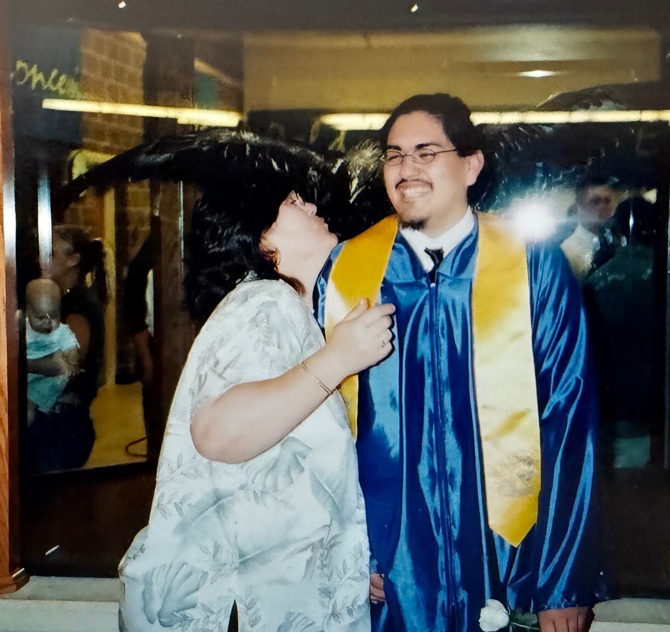 Graduate in a blue gown with a yellow stole smiles while embracing a person in a light dress at a celebration. This appears to be a graduation moment indoors with friends in the background.