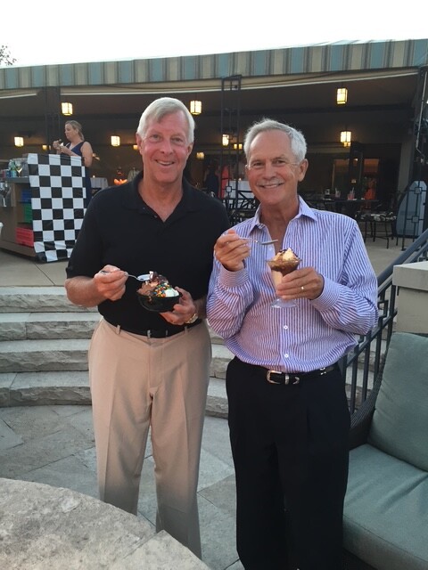 Two smiling men stand on a patio at an evening event, each holding a dessert cup; one in a black polo, the other in a striped shirt.