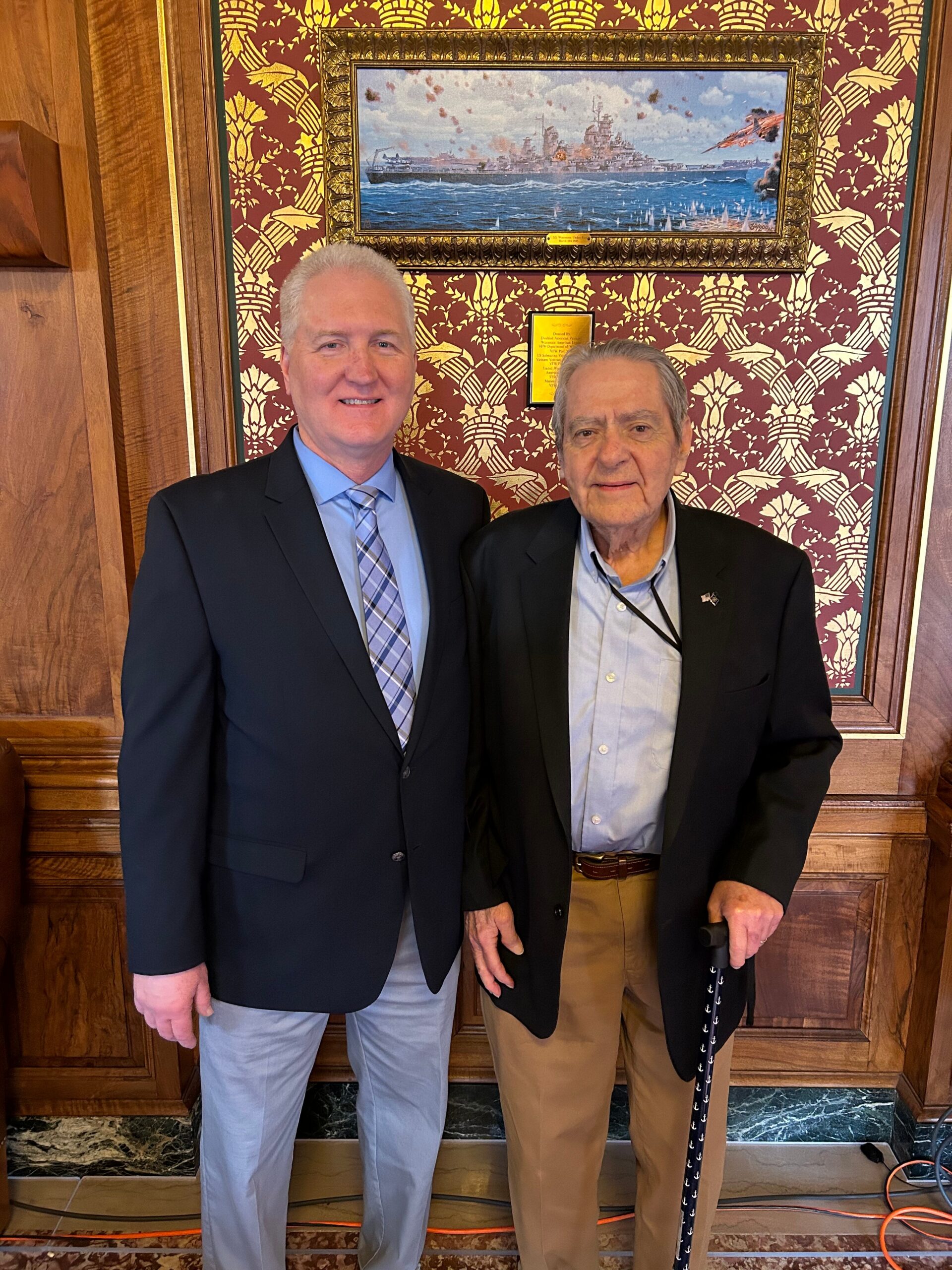 Two men in suits posing together in a wood-paneled hallway with ornate wallpaper and a framed painting behind them.