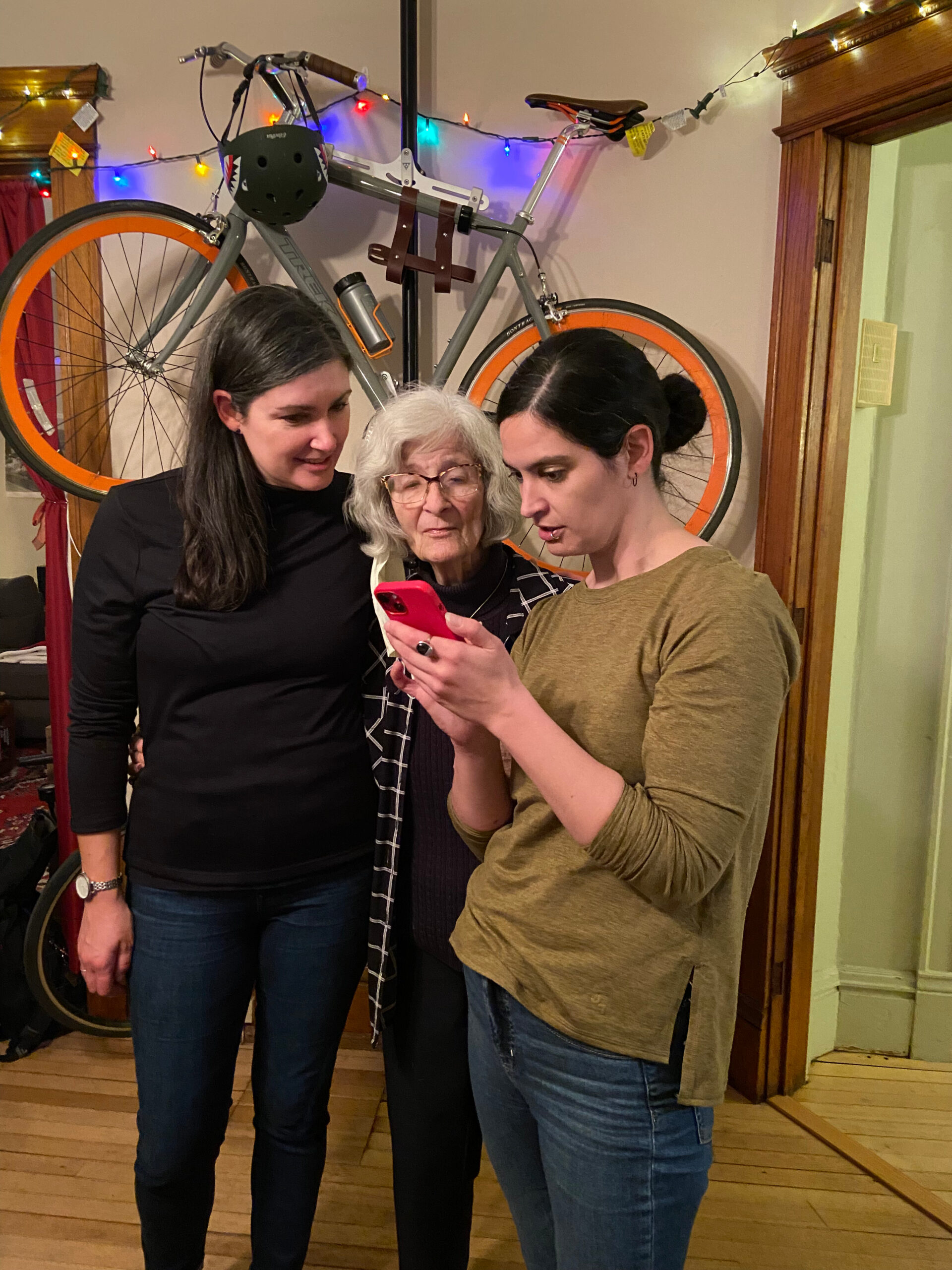 Three women gathered indoors, looking at a pink smartphone, with a wall-mounted bicycle and colorful lights in the background.