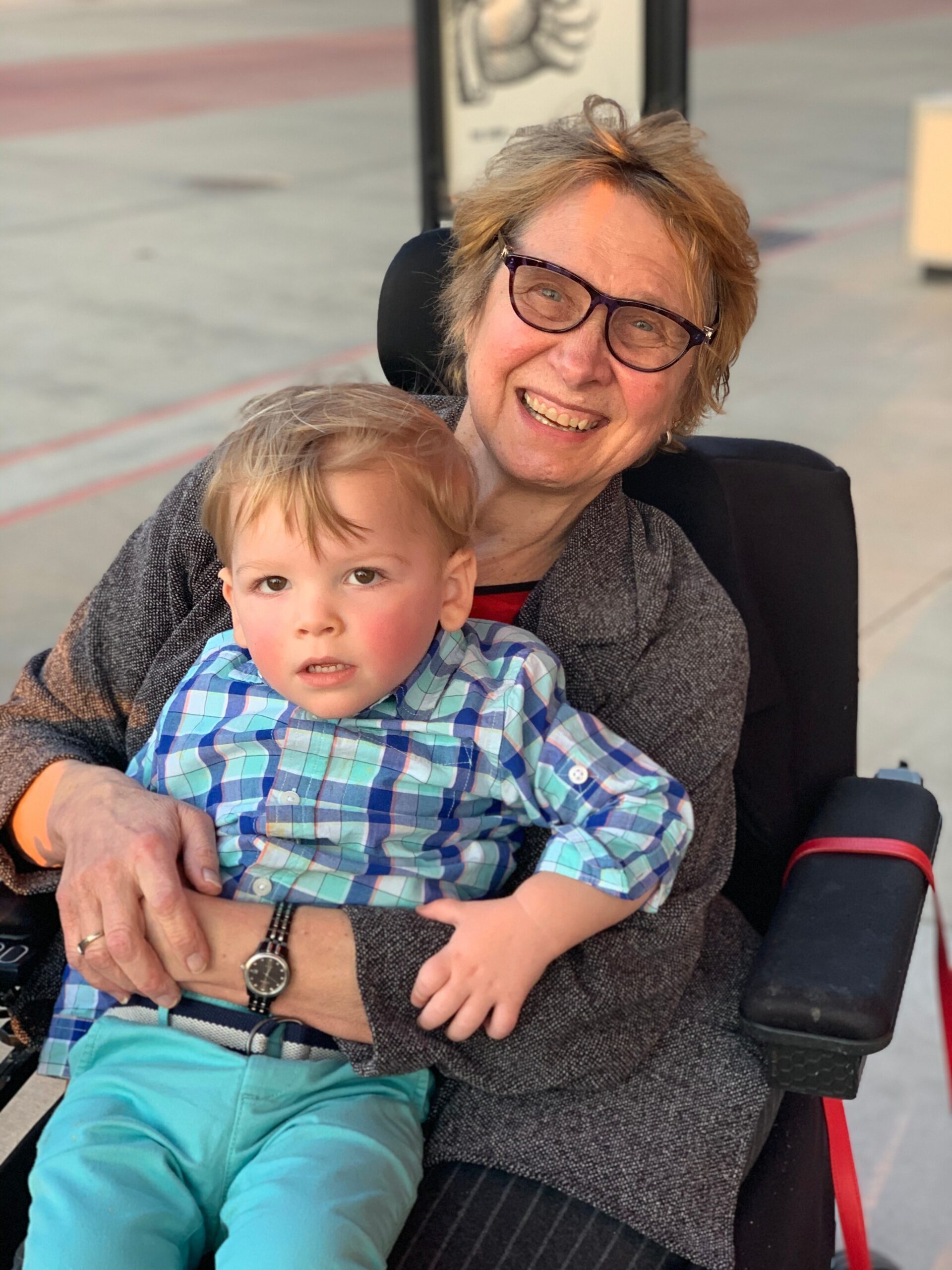 Smiling older woman with short hair and purple glasses sits in a wheelchair, holding a young child in a blue plaid shirt.