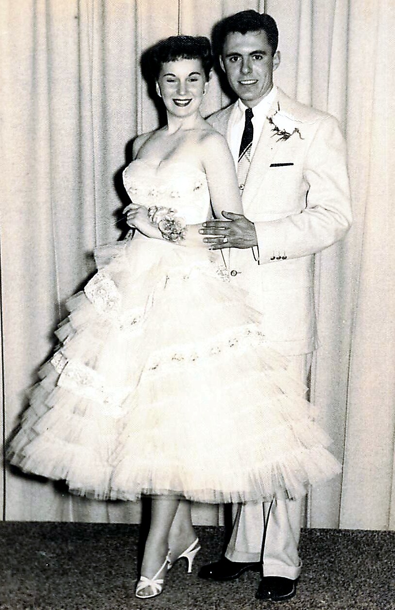 Bride in a strapless white wedding gown with layered tulle, standing beside a groom in a light suit and boutonniere, posing indoors.