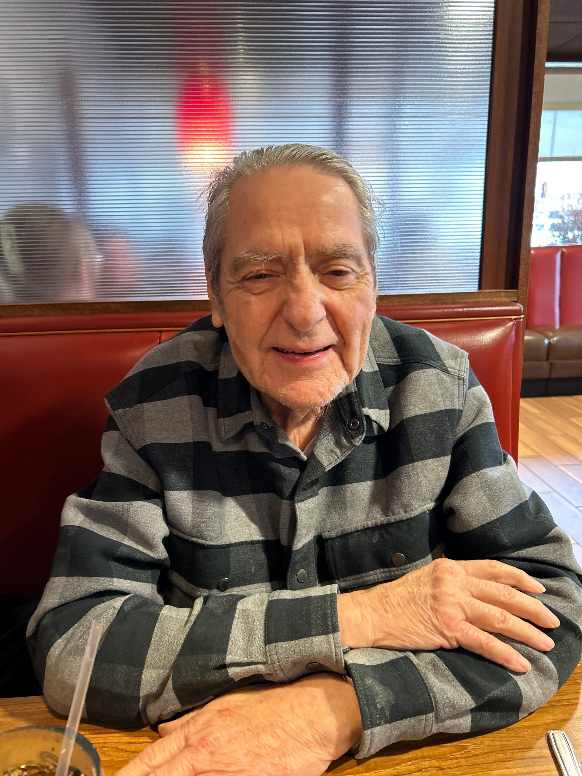Older man with gray hair in a black-and-gray striped shirt sits at a booth in a restaurant, arms crossed on a wooden table, beverage with a straw nearby.