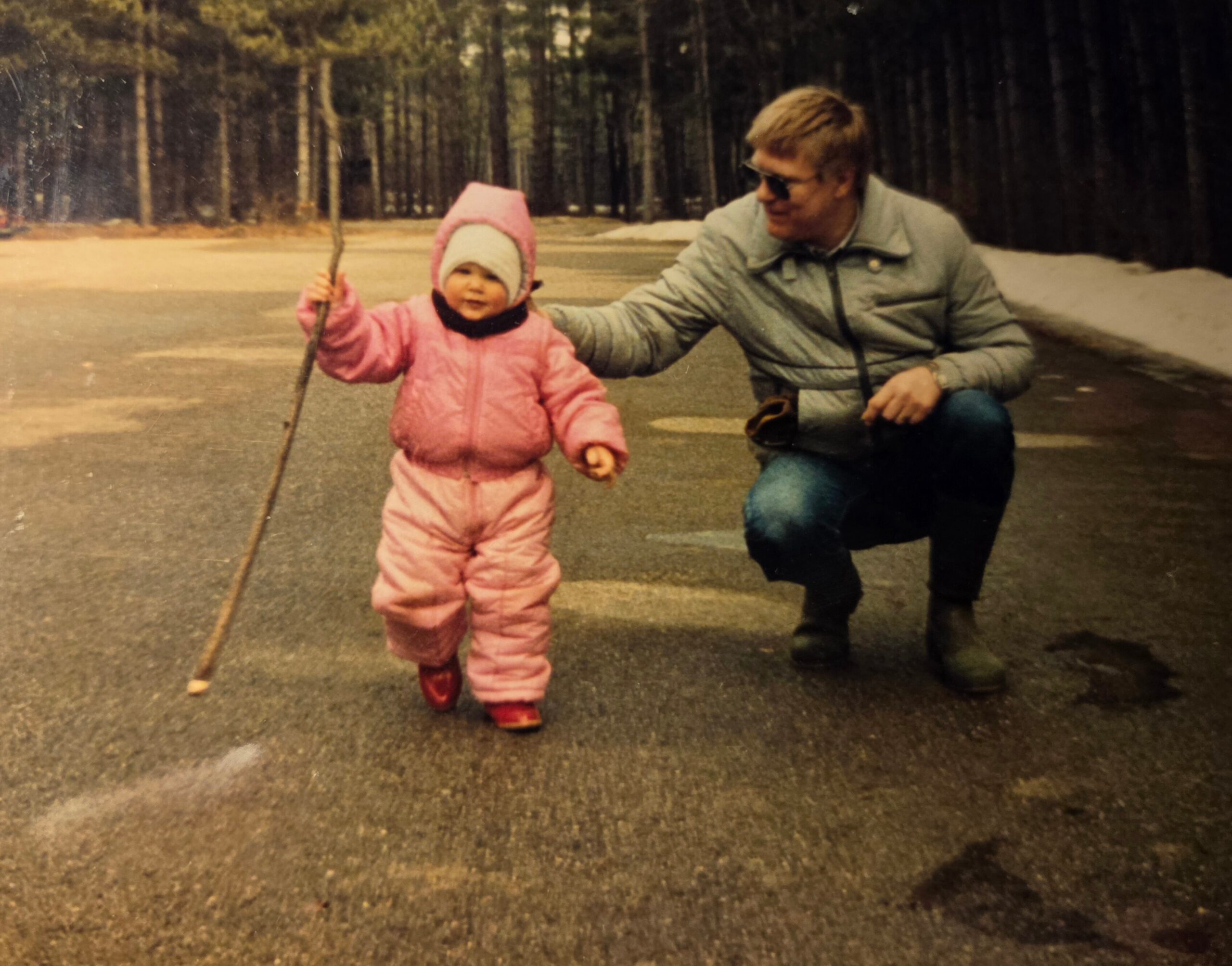 Toddler in a pink snowsuit walks with a stick while an adult man crouches beside her outdoors, holding her hand on a paved path with trees in the background.