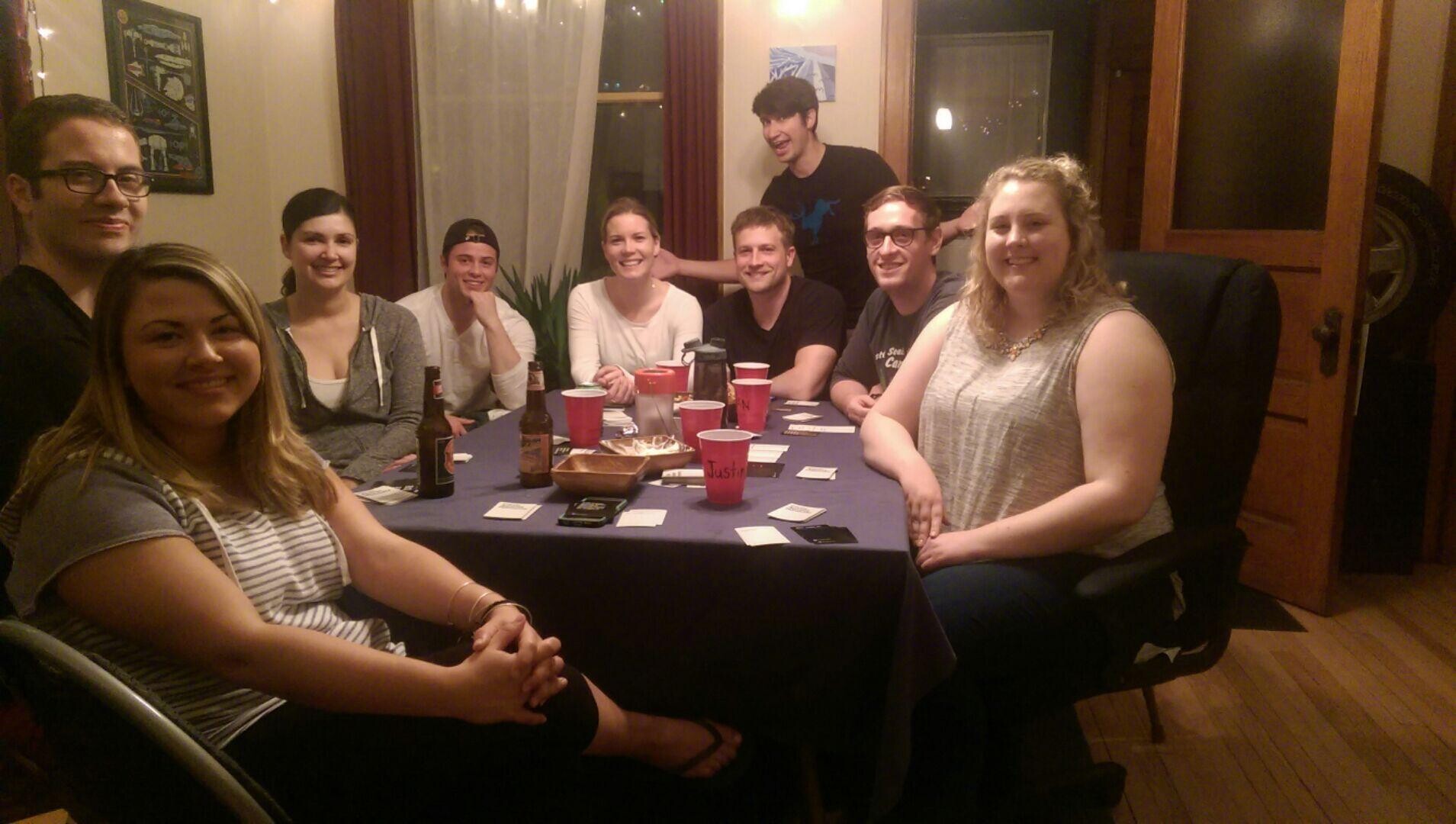 Nine friends sit around a dining table in a cozy living room, smiling, with red cups, beer bottles and cards scattered on the table.