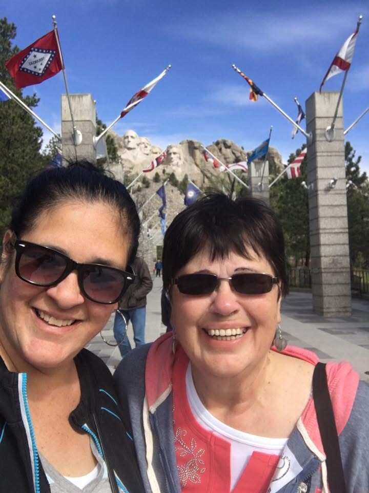 Two women wearing sunglasses take a smiling selfie with Mount Rushmore and flagpoles in the background.