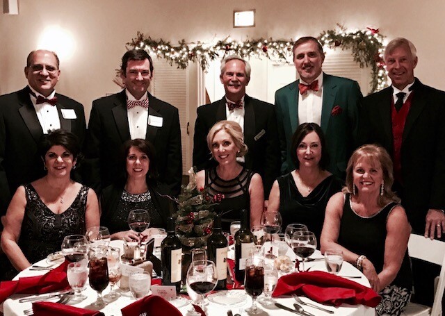 Ten adults in formal attire (five women seated, five men standing) posing around a decorated holiday dinner table with a small Christmas tree centerpiece and garland lights in the background.