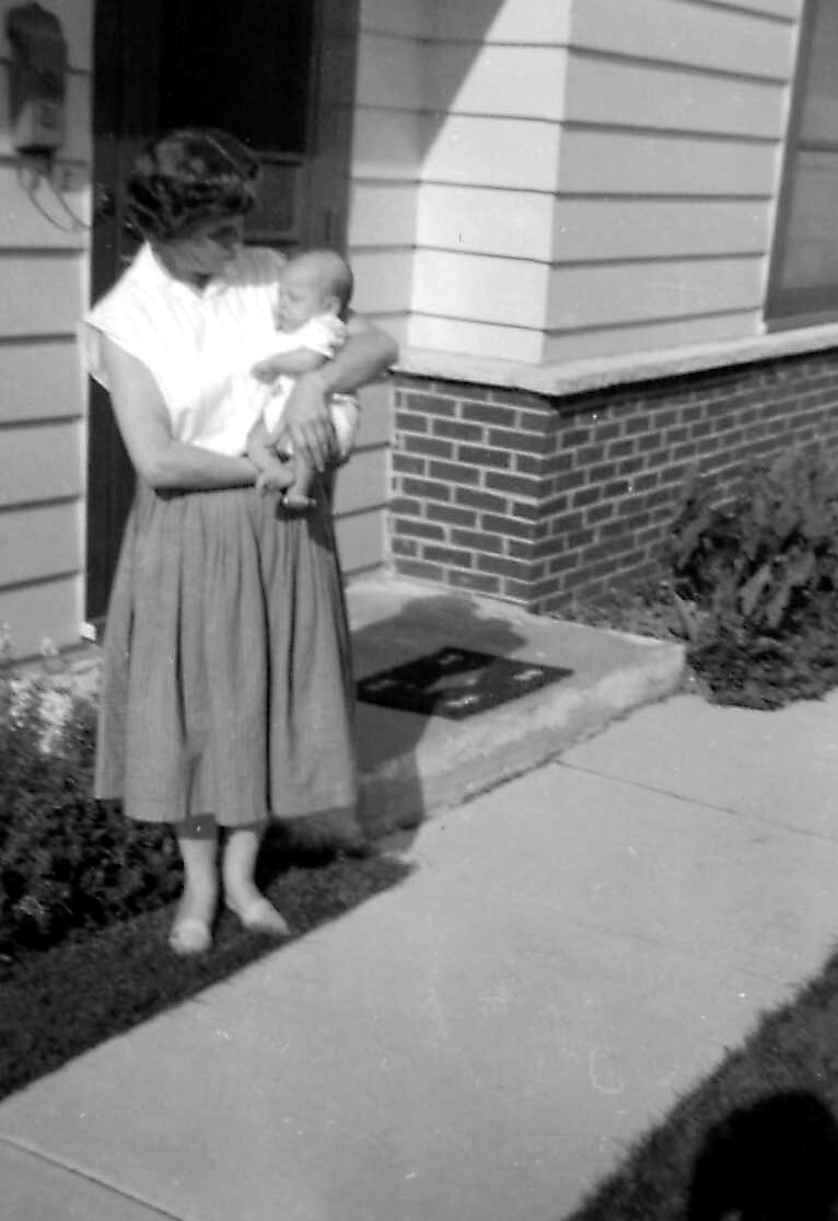 Woman wearing a white blouse and knee-length skirt stands on a sidewalk outside a house, cradling a baby in her arms.