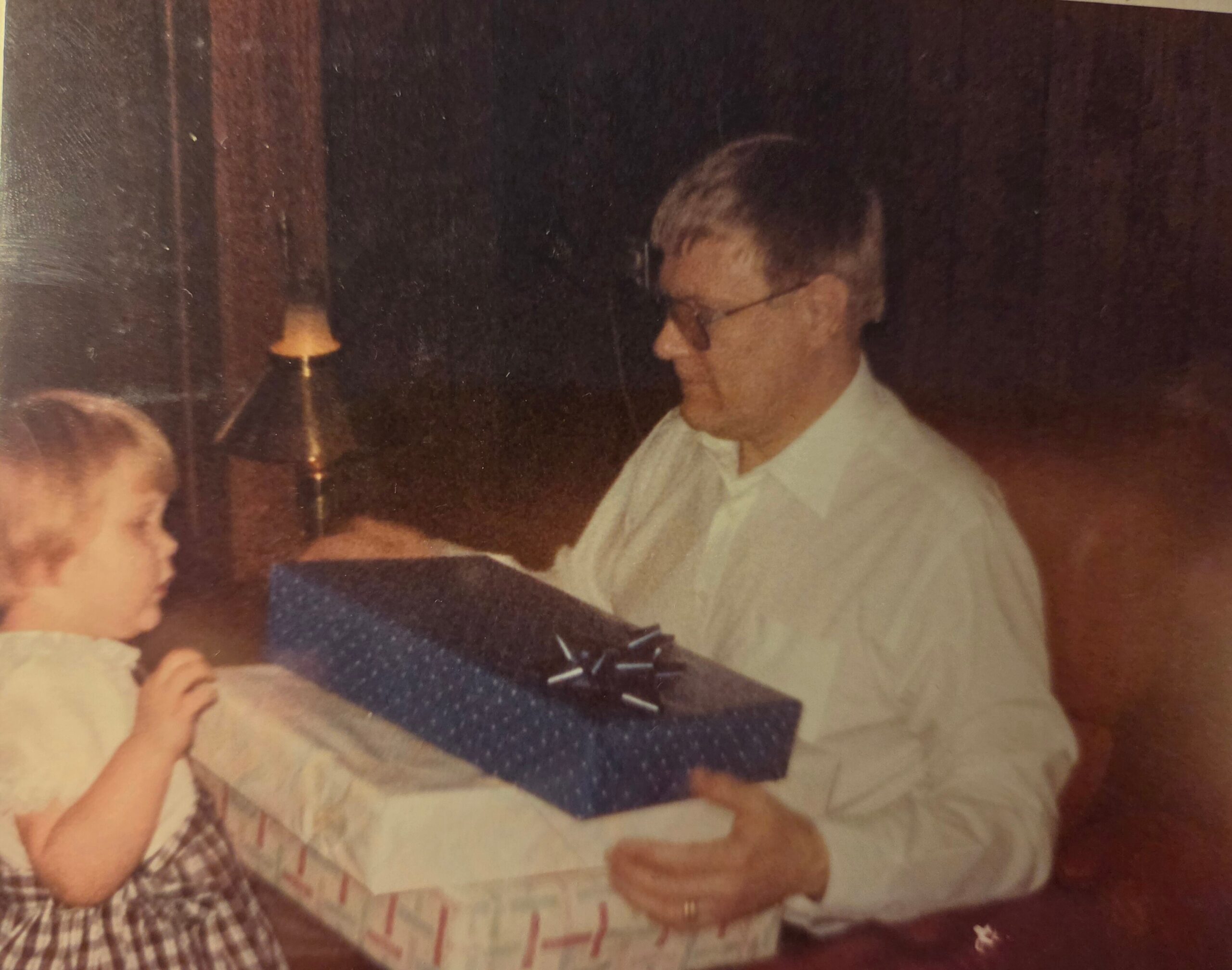 Side profile of a man in a white shirt and tie, wearing glasses, looking to the right at a family gathering; a baby sits at a table in the foreground.