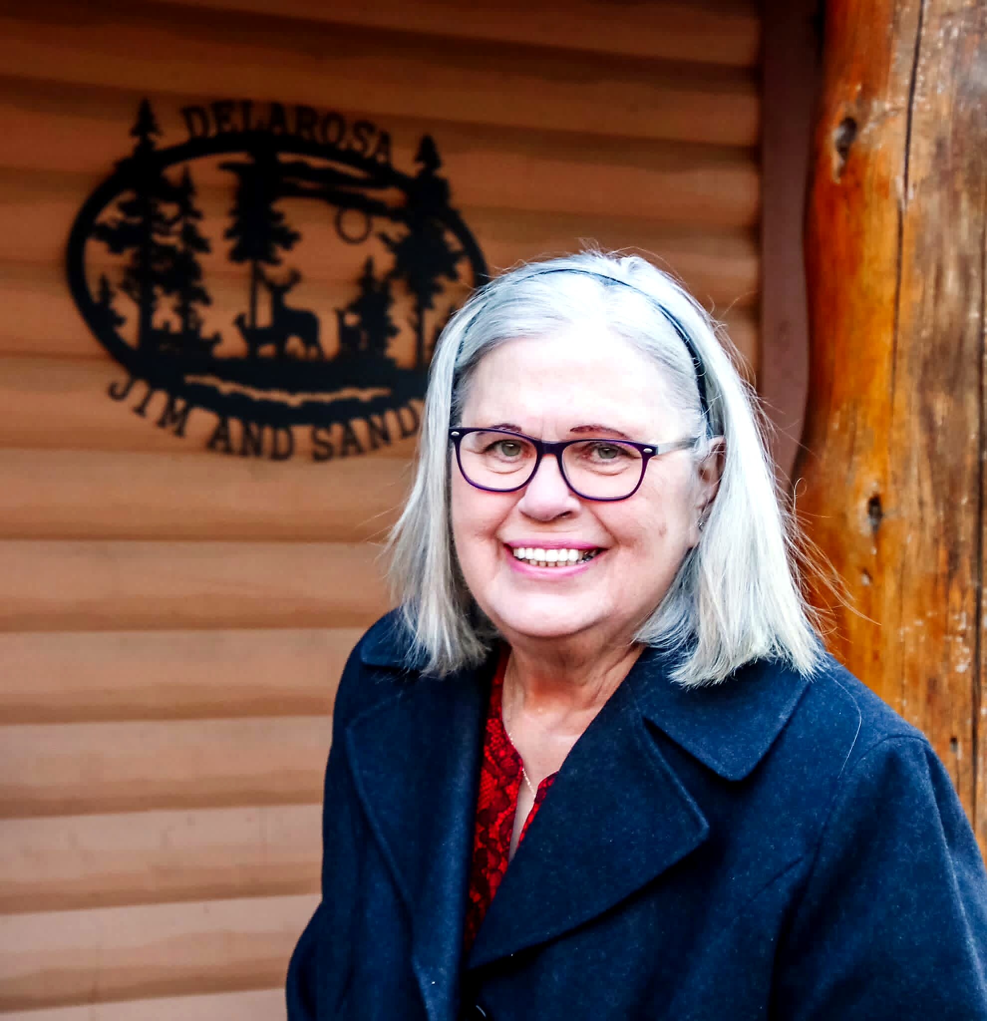 Portrait of a smiling woman with light gray hair and purple glasses, wearing a navy blazer in front of a wooden wall with a logo behind her.