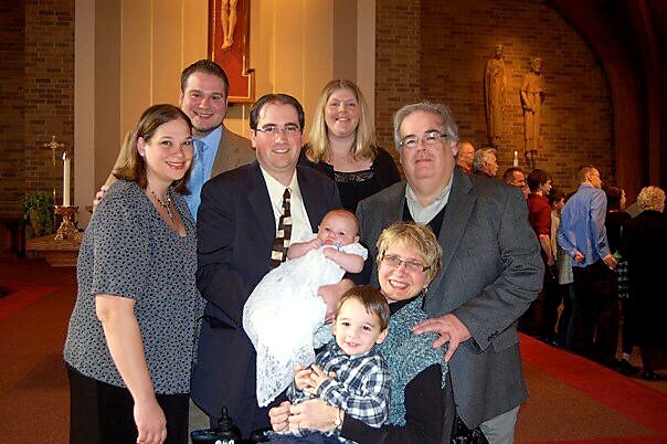 Group of eight adults and two children posing for a photo in a church sanctuary, including a baby in a white blanket and a smiling toddler at front.