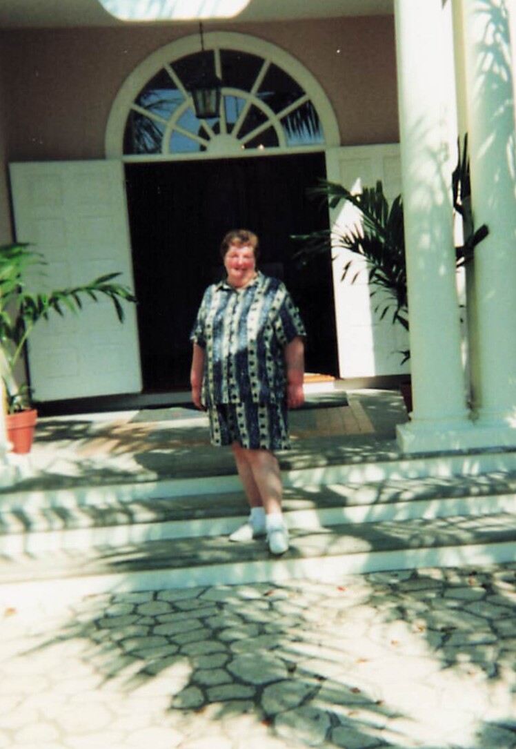 Older woman standing on front steps in a blue and white patterned dress, arched doorway behind her.