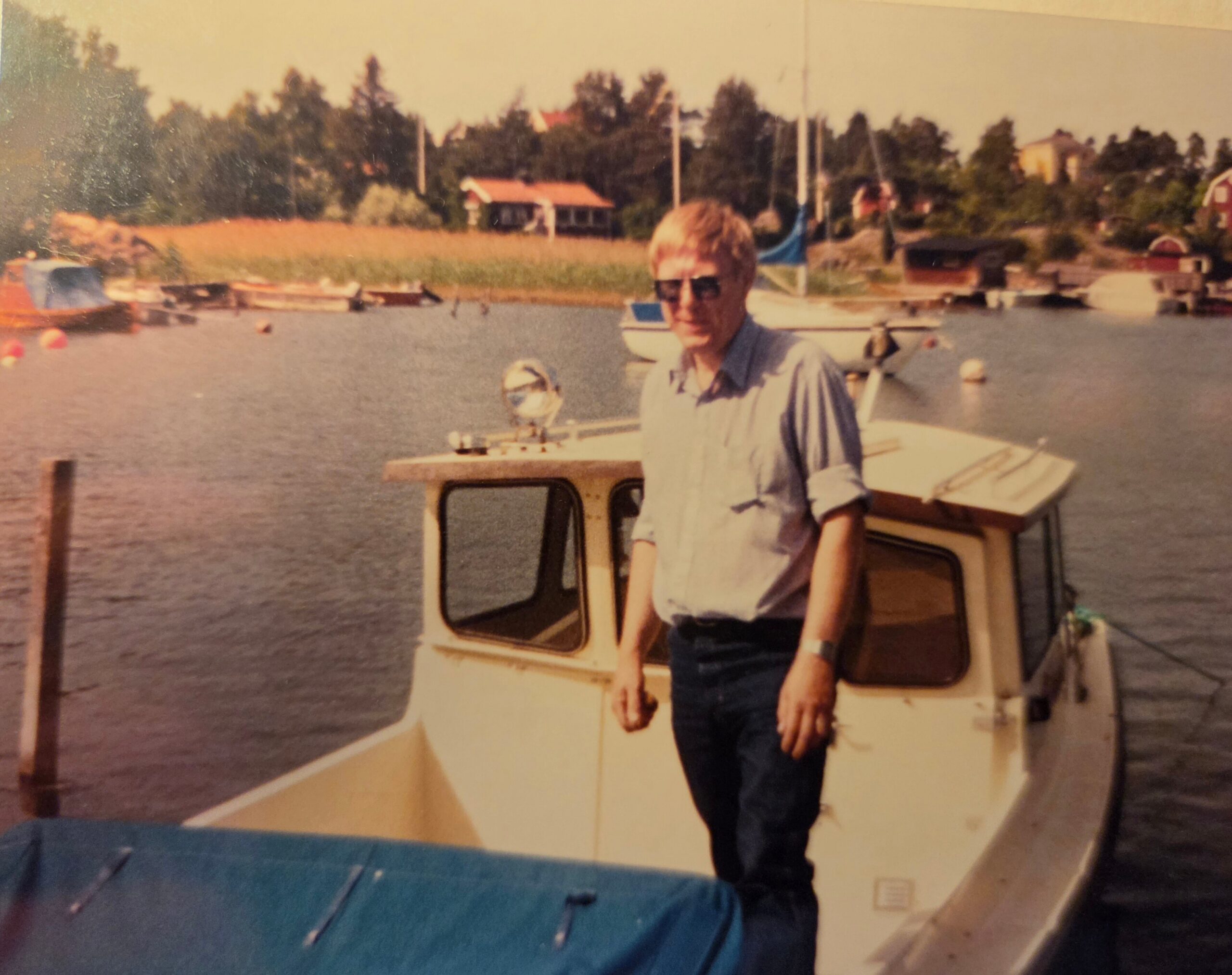 Man in sunglasses standing on the deck of a small boat at a marina, with water and distant houses in the background, a sunny day.