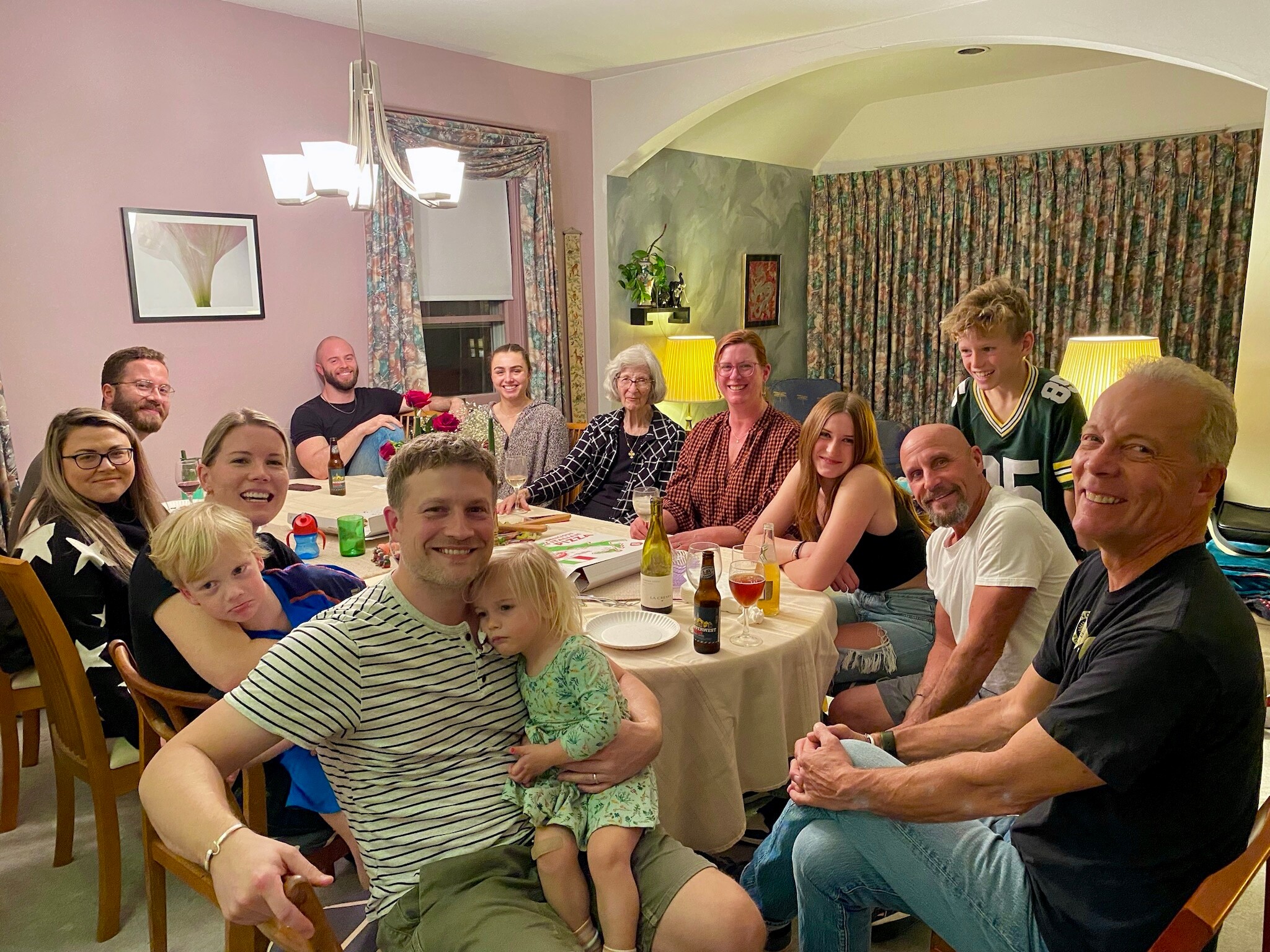 Family and friends gathered around a dining table in a pink-walled living room, smiling for a group photo during a celebration.