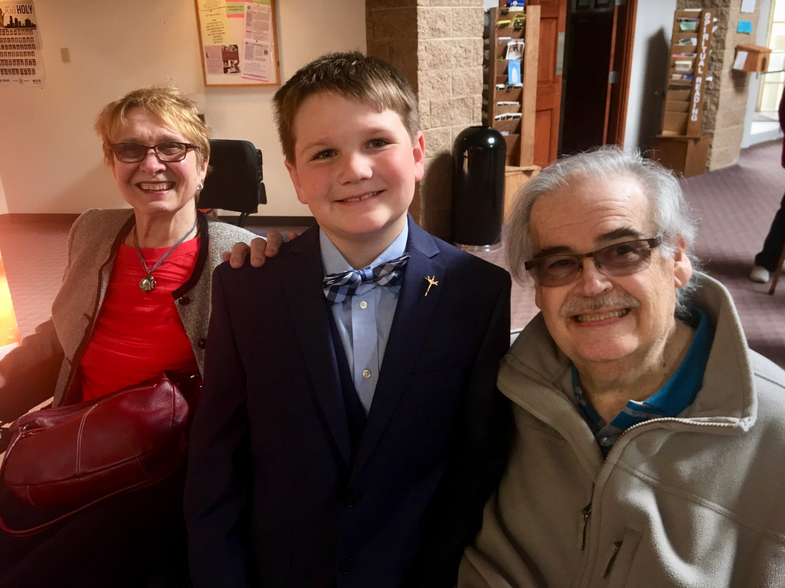 Three smiling people posing in a building lobby: a woman with short blond hair and glasses, a boy in a suit, and an older man with gray hair and glasses.