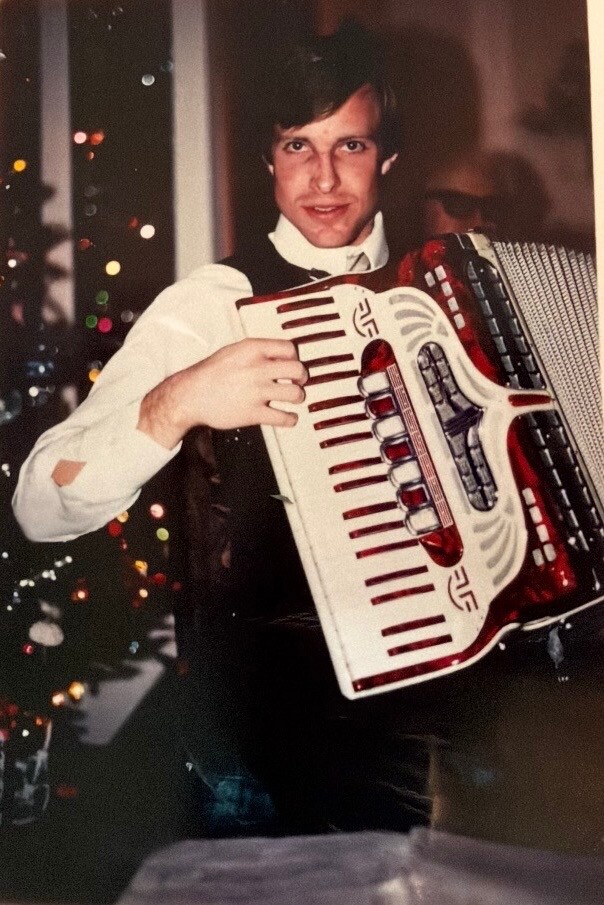 Man in a white shirt and vest playing a red-and-white accordion indoors with Christmas tree lights in the background.