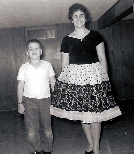 Woman in a black top and patterned lace skirt stands beside a young boy in a white polo shirt indoors, both smiling.
