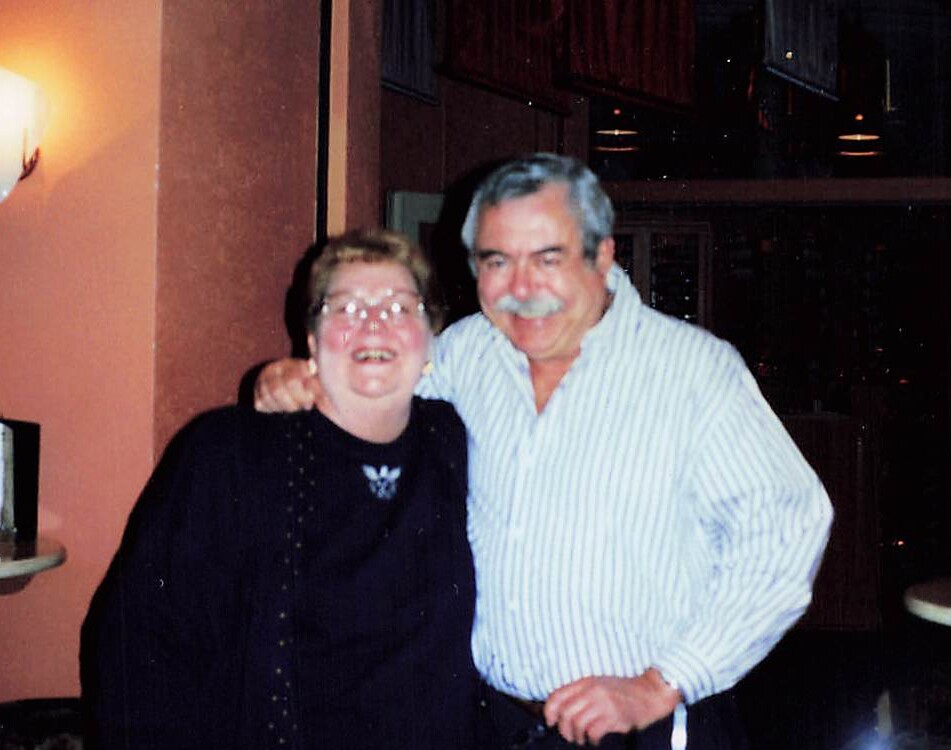 Two smiling older adults pose together with arms around each other at a social gathering, indoors near a warm light.