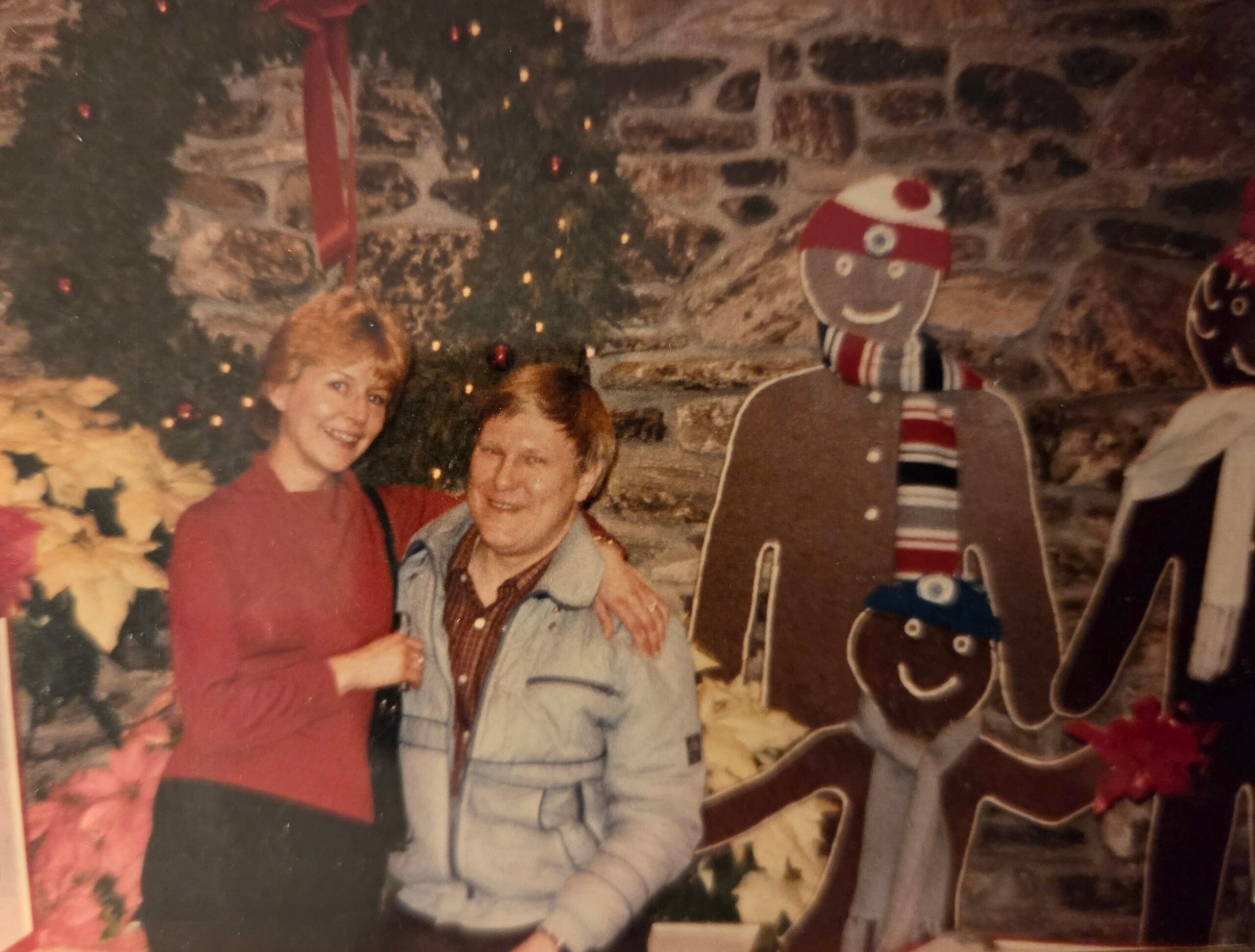 Two people posing for a holiday photo in a living room, with a Christmas tree, stockings, and festive decorations in the background.