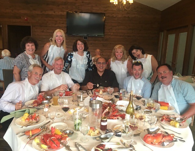 Twelve adults wearing bibs around a round table with lobsters and wine in a wood-paneled restaurant, smiling for a group photo.
