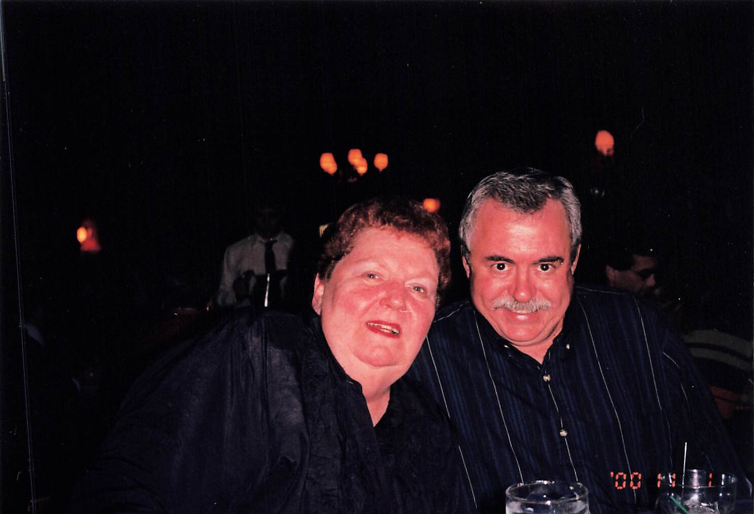 Smiling couple seated at a dimly lit gathering, posing for the camera with drinks nearby.