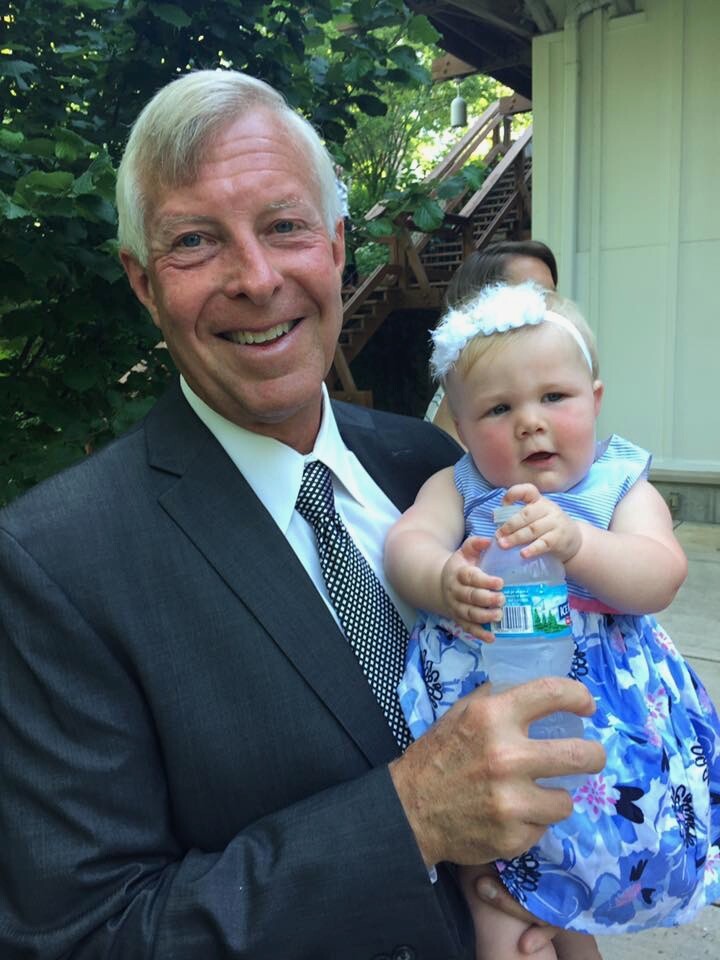 Smiling man in a suit holding a baby girl in a blue floral dress outdoors with greenery behind them.