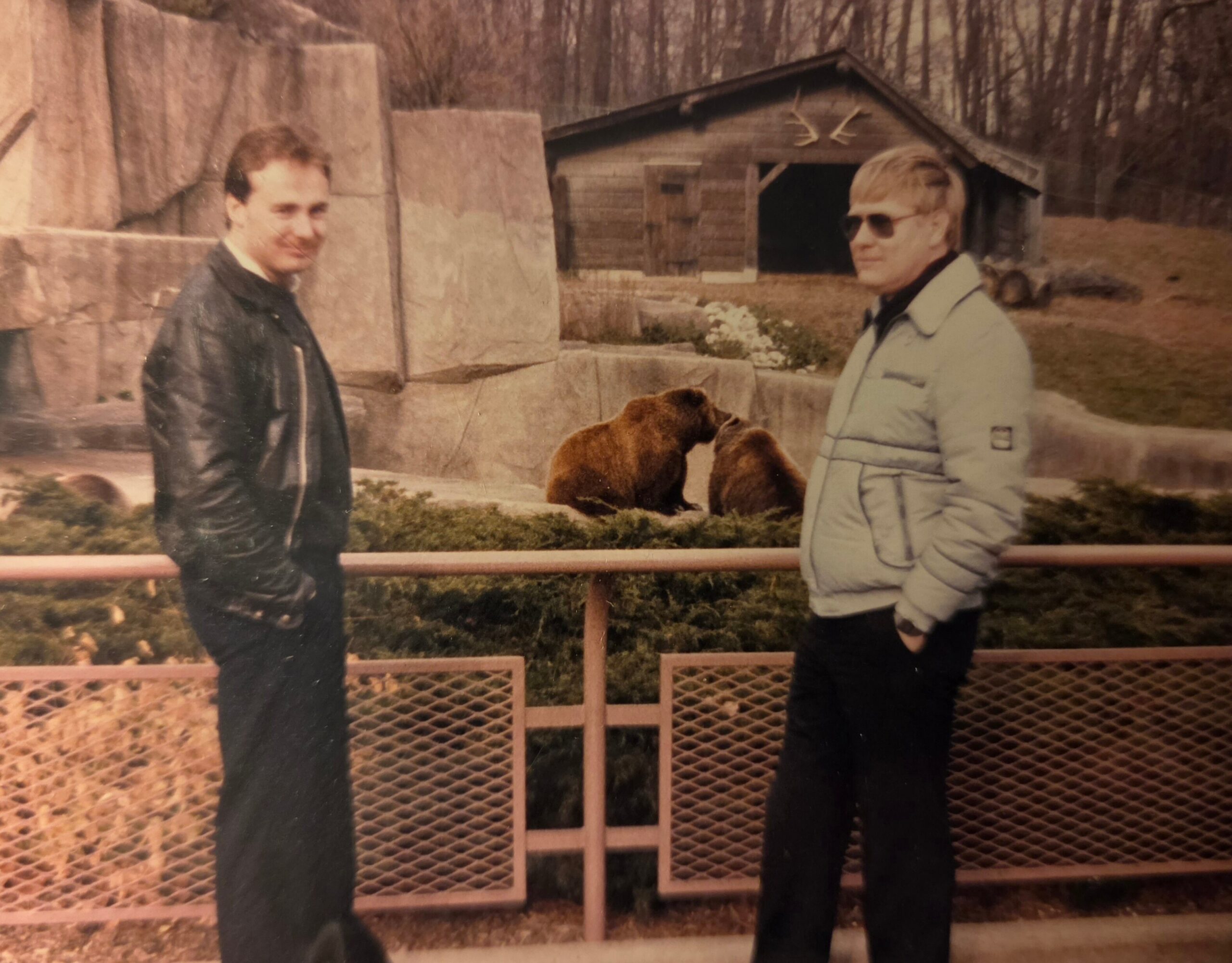 Two men in jackets stand apart outdoors beside a brown bear sitting between them, with a wooden shed and trees in the background.