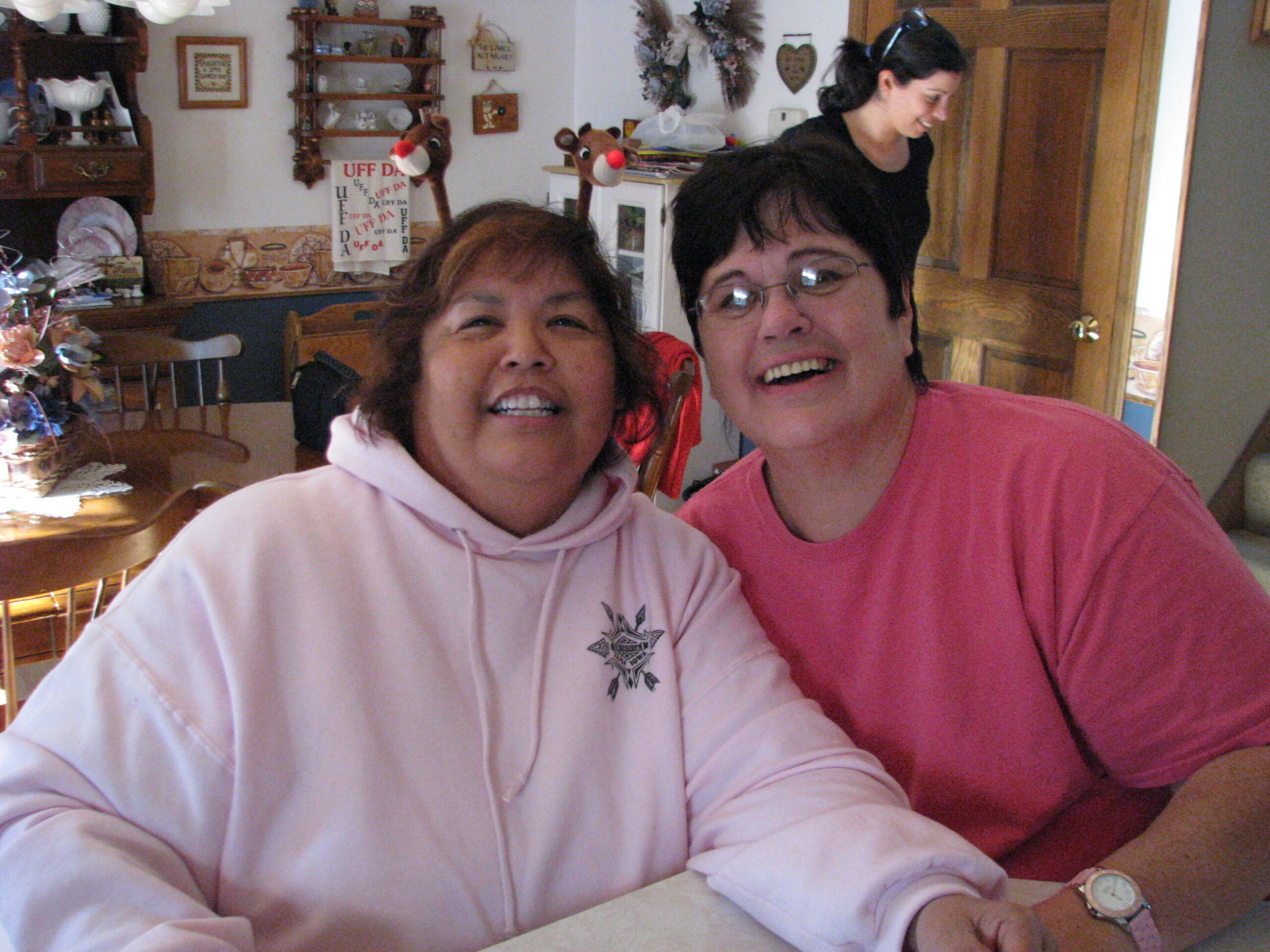 Two women smiling for a selfie in a cozy kitchen/dining area with decorative shelves and holiday ornaments in the background.