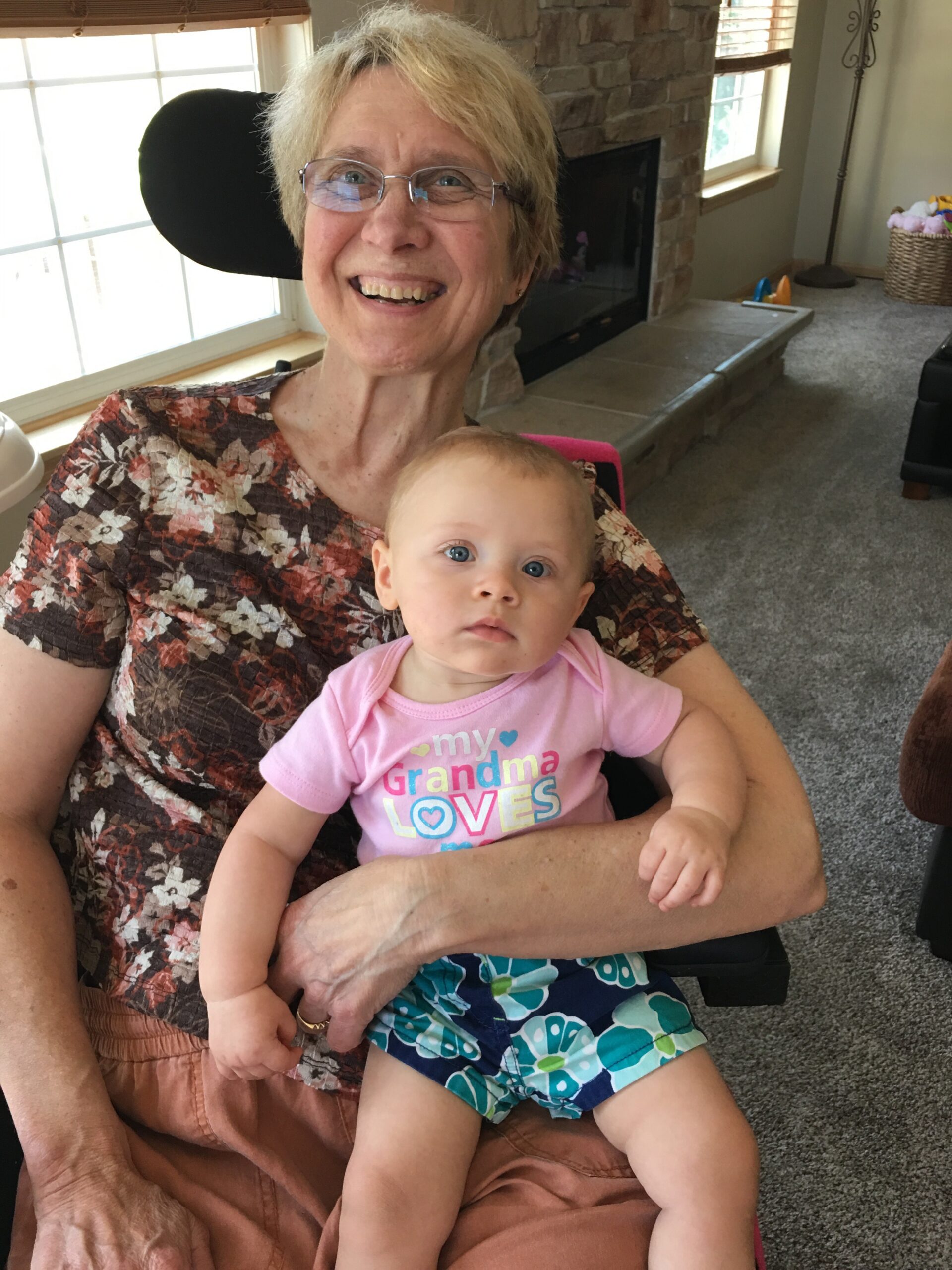 Smiling grandmother with short blonde hair and glasses sits indoors, holding a baby girl in a pink onesie with the text 'my Grandma loves' visible.