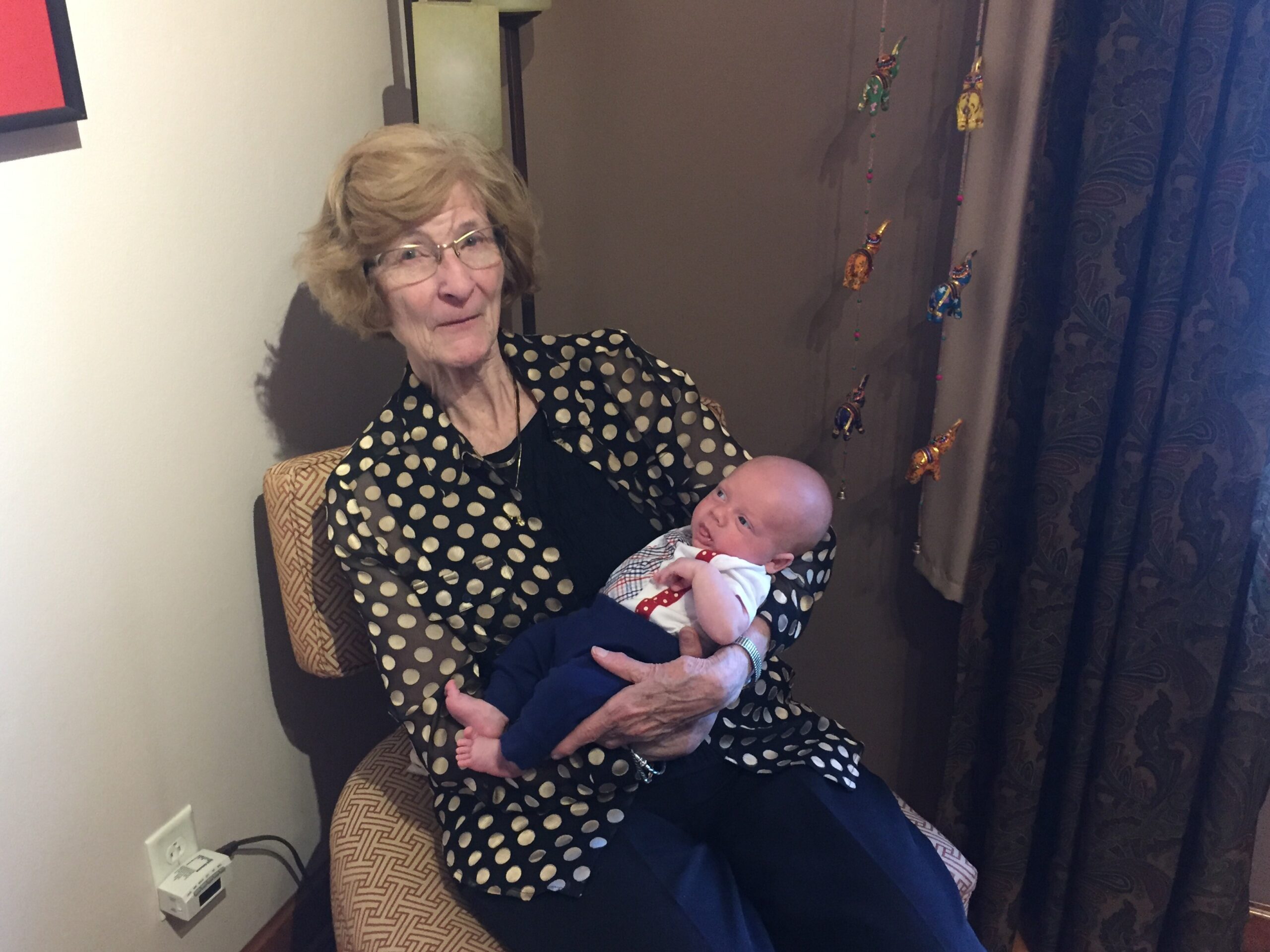 Older woman in a polka-dot blouse holding a baby in her arms on a patterned chair indoors.