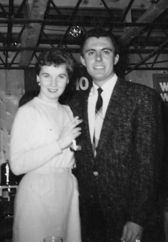 A smiling couple posing together at a social event, the woman in a light dress and the man in a dark suit and tie, indoors at a party.