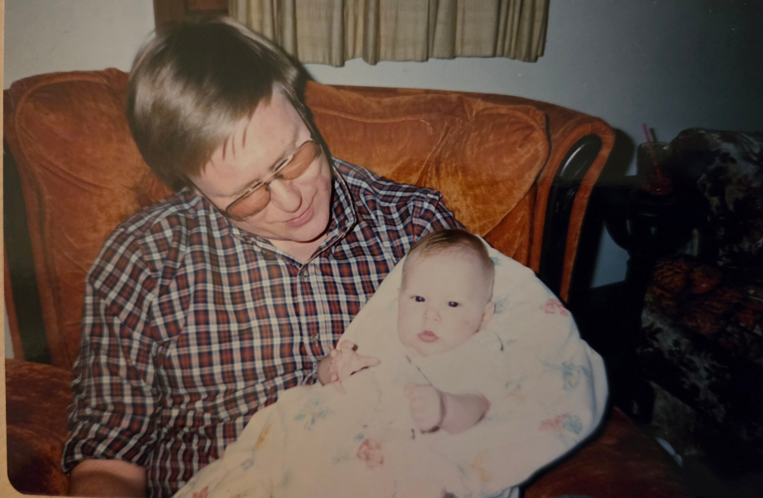 Man wearing plaid shirt and glasses cradles a newborn wrapped in a blanket on an orange couch in a living room.