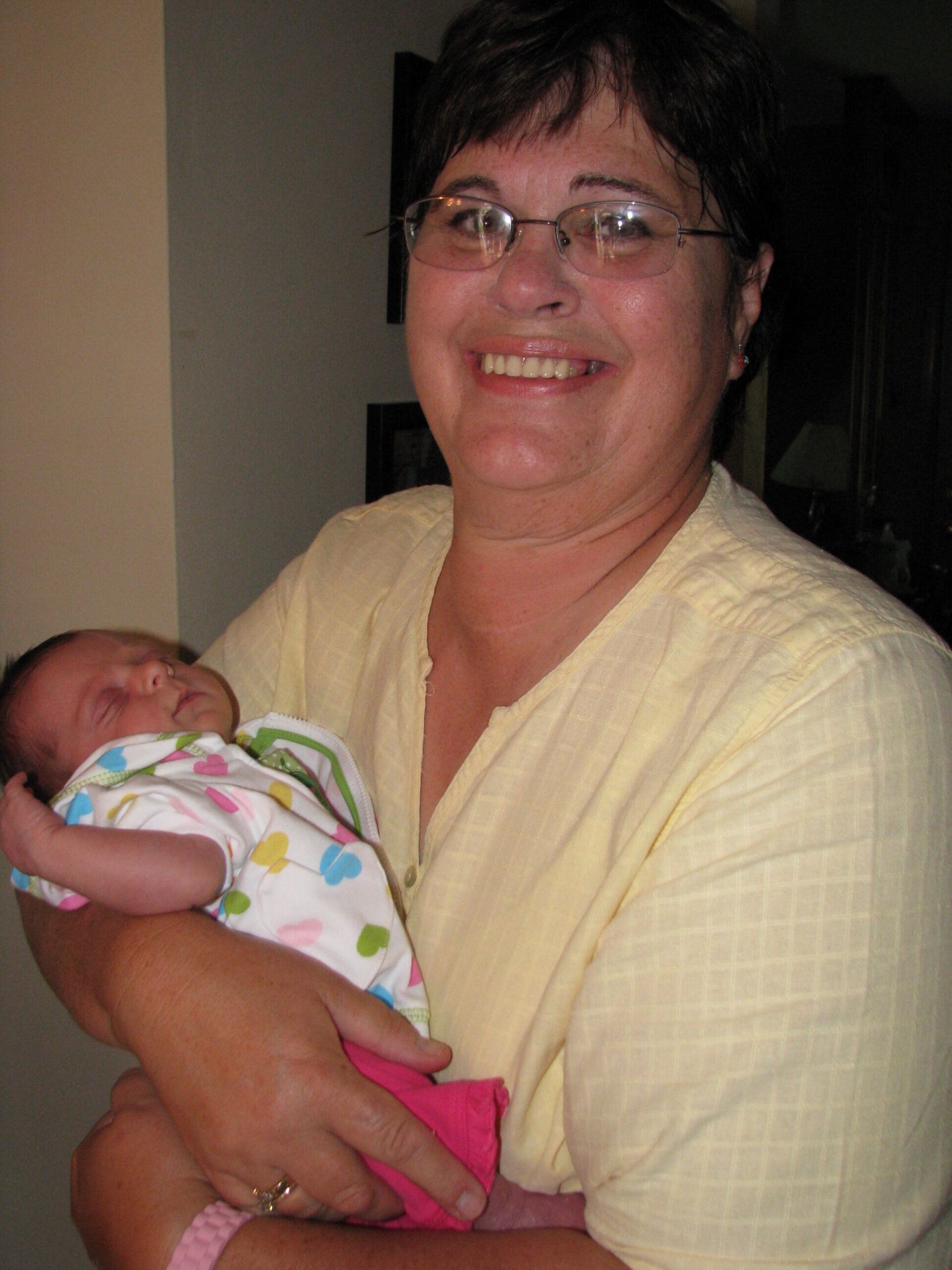 Smiling woman wearing glasses holds a newborn baby wrapped in a colorful patterned blanket.